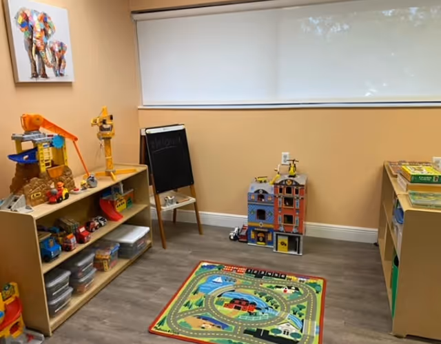 Colorful children's play area inside a senior facility, featuring toy houses, a rug, and shelves.