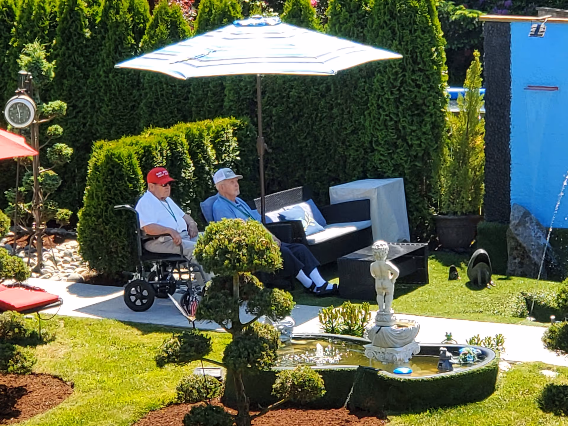 Two elderly men sitting outdoors in a garden area. One man is in a wheelchair wearing a red cap, white shirt, and beige pants. The other man is seated on a black outdoor sofa under a large white umbrella, wearing a light blue shirt and dark pants. The garden features neatly trimmed bushes, a small decorative fountain with a statue, and a blue wall with a water feature in the background.
