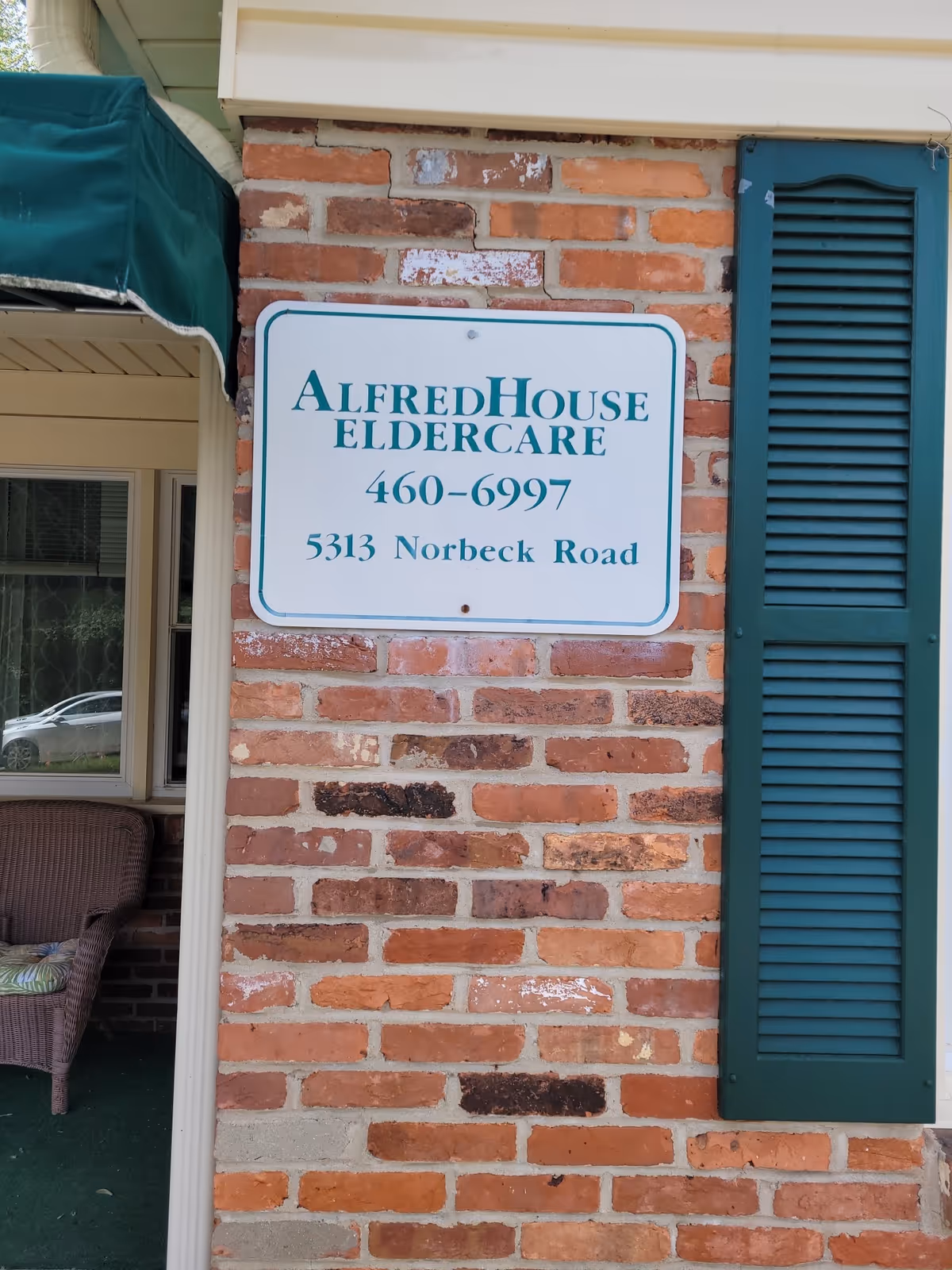 Exterior brick wall with a sign for AlfredHouse ElderCare and a green shutter.