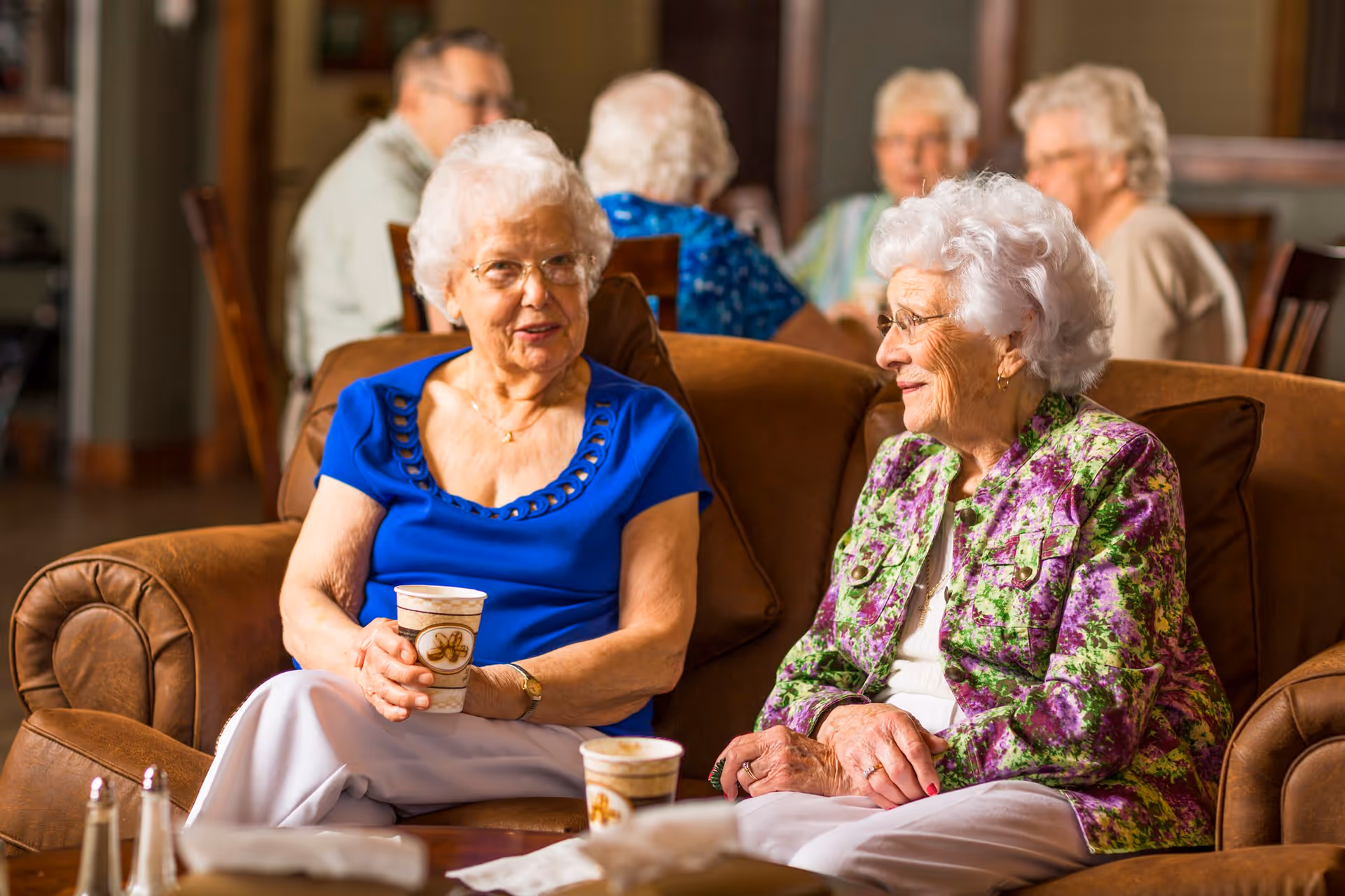 Two elderly women sitting on a brown leather couch in a communal living area, engaged in conversation while holding disposable coffee cups. In the background, other elderly individuals are seated and interacting around tables.