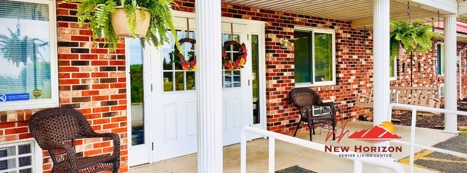 Front entrance of a brick senior living facility with white doors, hanging plants, and porch seating.