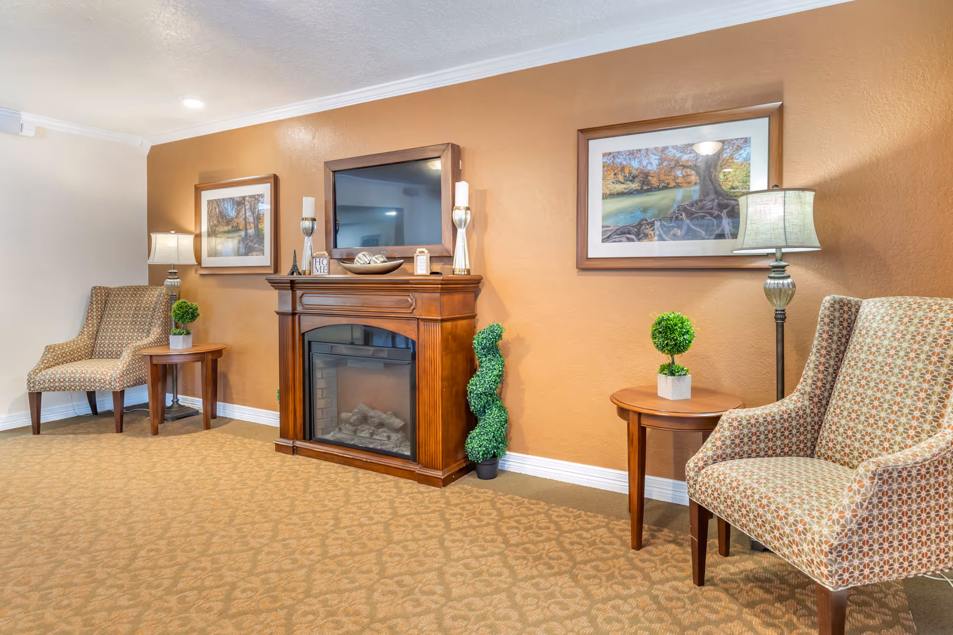 Cozy senior living lounge with fireplace, TV, two patterned chairs, and side tables.