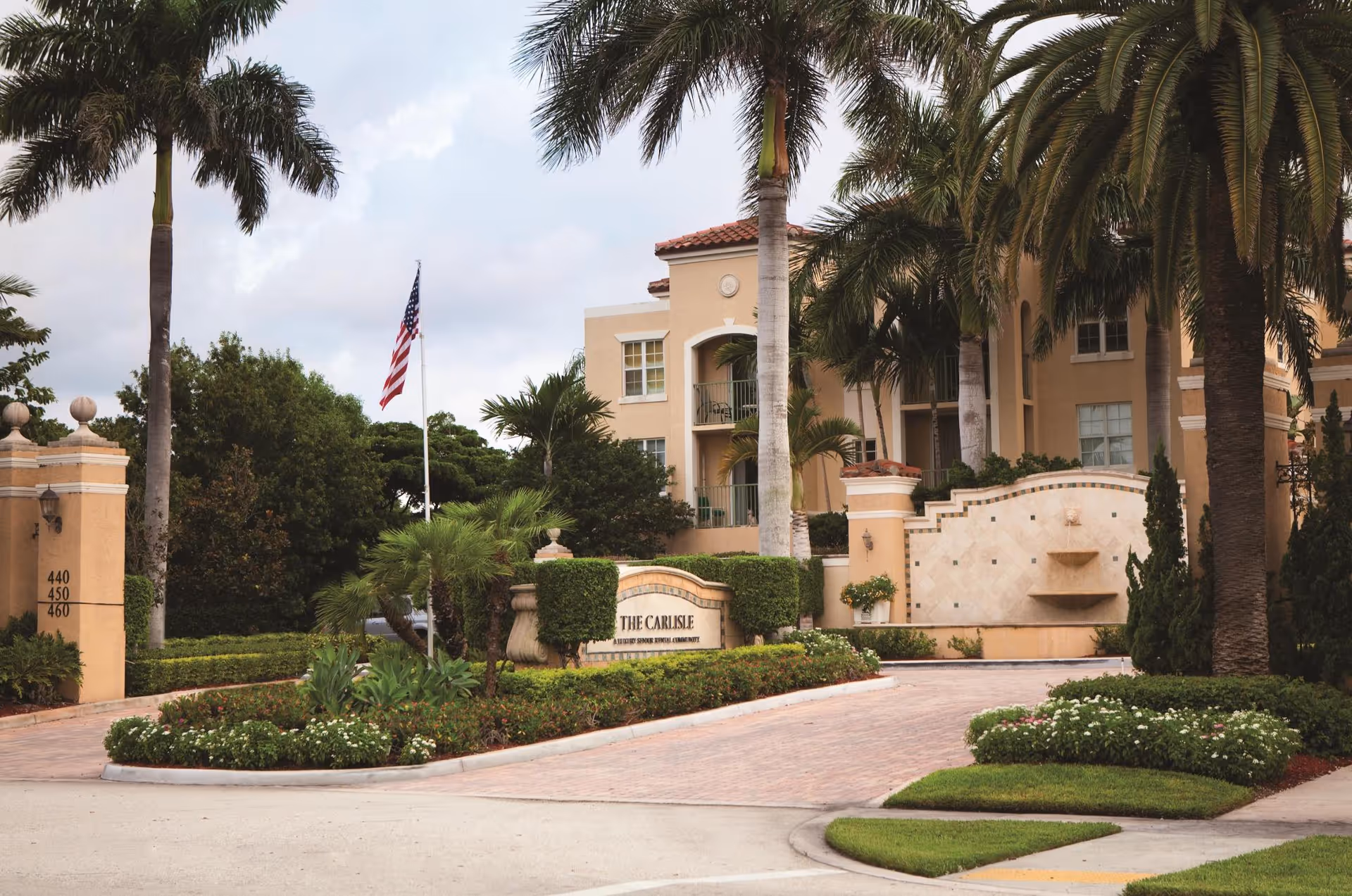 Entrance to The Residences at The Carlisle Palm Beach featuring a landscaped driveway with palm trees, an American flag on a flagpole, and a beige building with balconies in the background.
