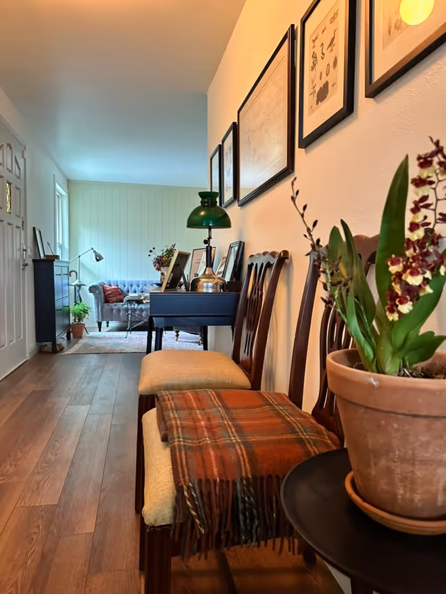 Narrow hallway leads to a cozy living area with chairs, desk, lamps, and plants.