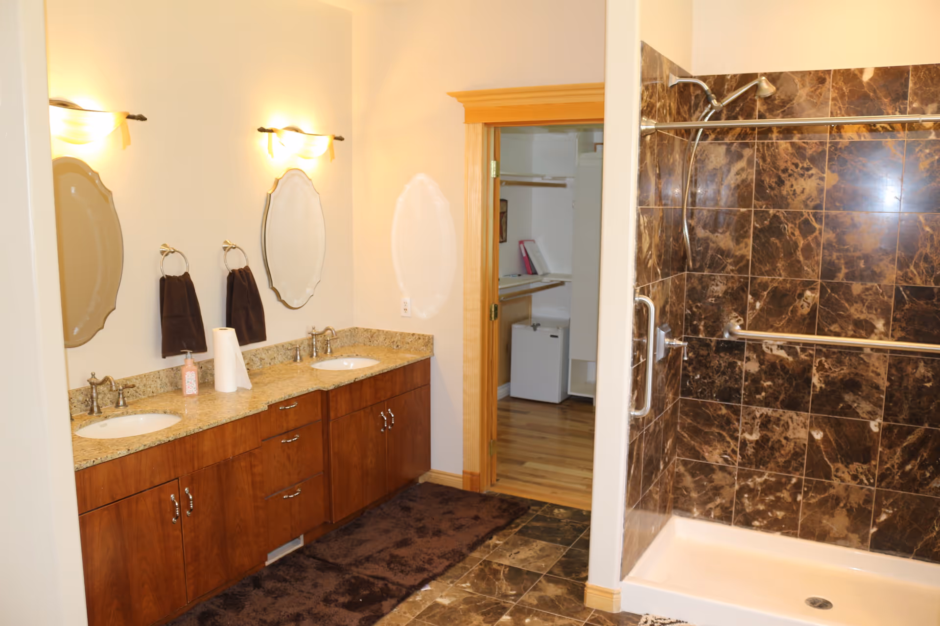 A bathroom with double sinks, granite counters, mirrors, dark-tiled shower, and doorway to a closet.