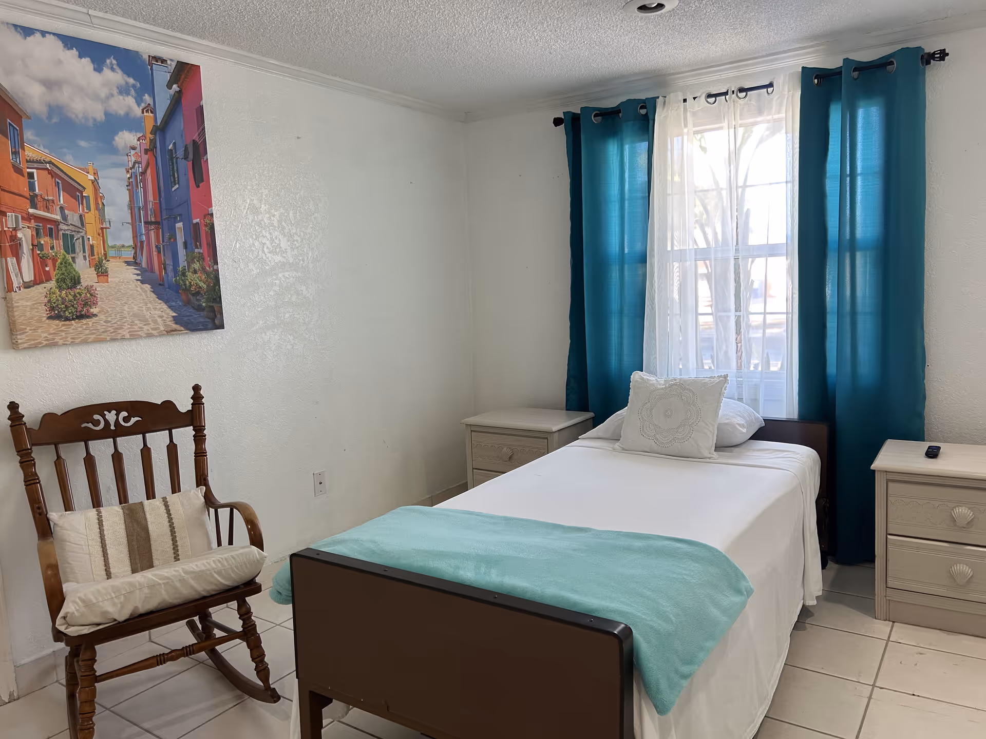 A furnished bedroom with a single bed, nightstands, teal curtains, a rocking chair, and wall art.