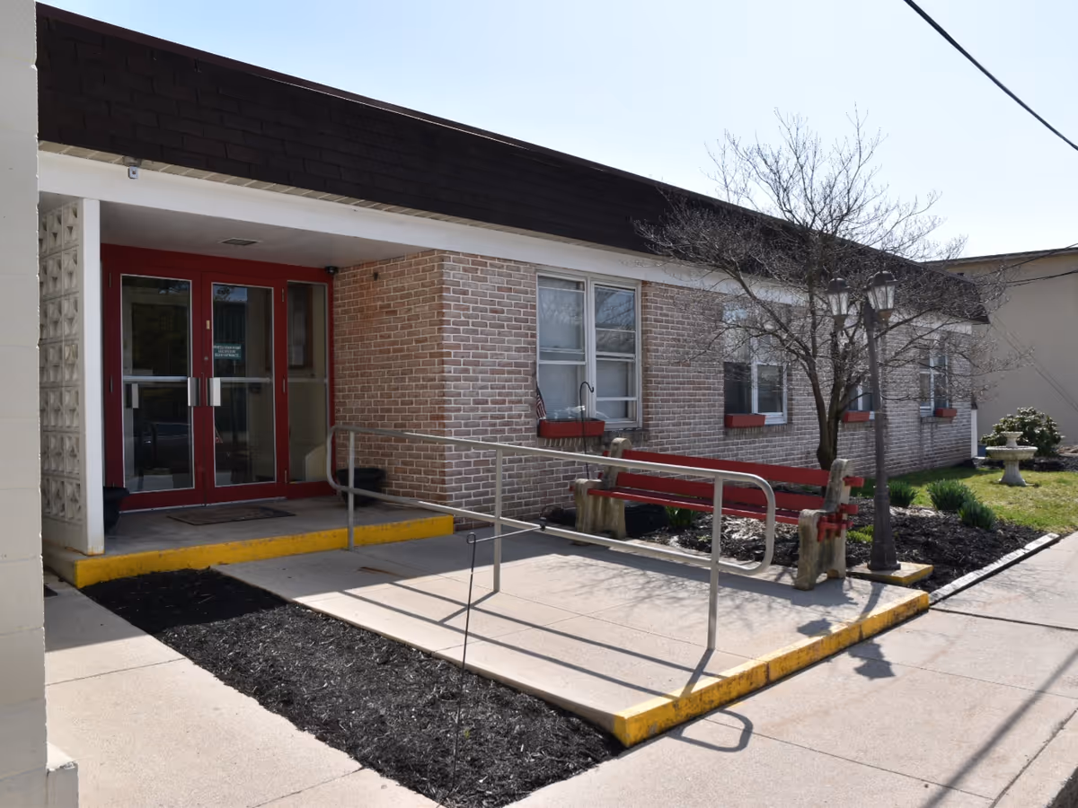 Exterior view of a single-story brick building with a red-framed glass double door entrance. There is a concrete ramp with metal handrails leading to the entrance, a red bench, a leafless tree, and a lamp post near the sidewalk. The area is landscaped with mulch and some greenery.