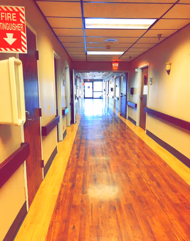 A long, well-lit hallway with wooden flooring and beige walls in a rehabilitation and healing facility. Several doors line both sides of the corridor, with handrails along the walls. A red fire extinguisher sign is visible on the left side near the ceiling, and an exit sign is illuminated at the far end of the hallway.