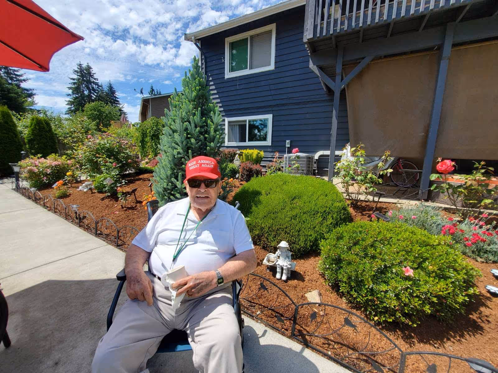 An elderly man wearing a red 'Make America Great Again' cap, sunglasses, a white polo shirt, and beige pants is sitting outdoors on a chair next to a garden with various green bushes and flowers. Behind him is a blue building with white-trimmed windows and a balcony. The sky is partly cloudy with blue patches visible.