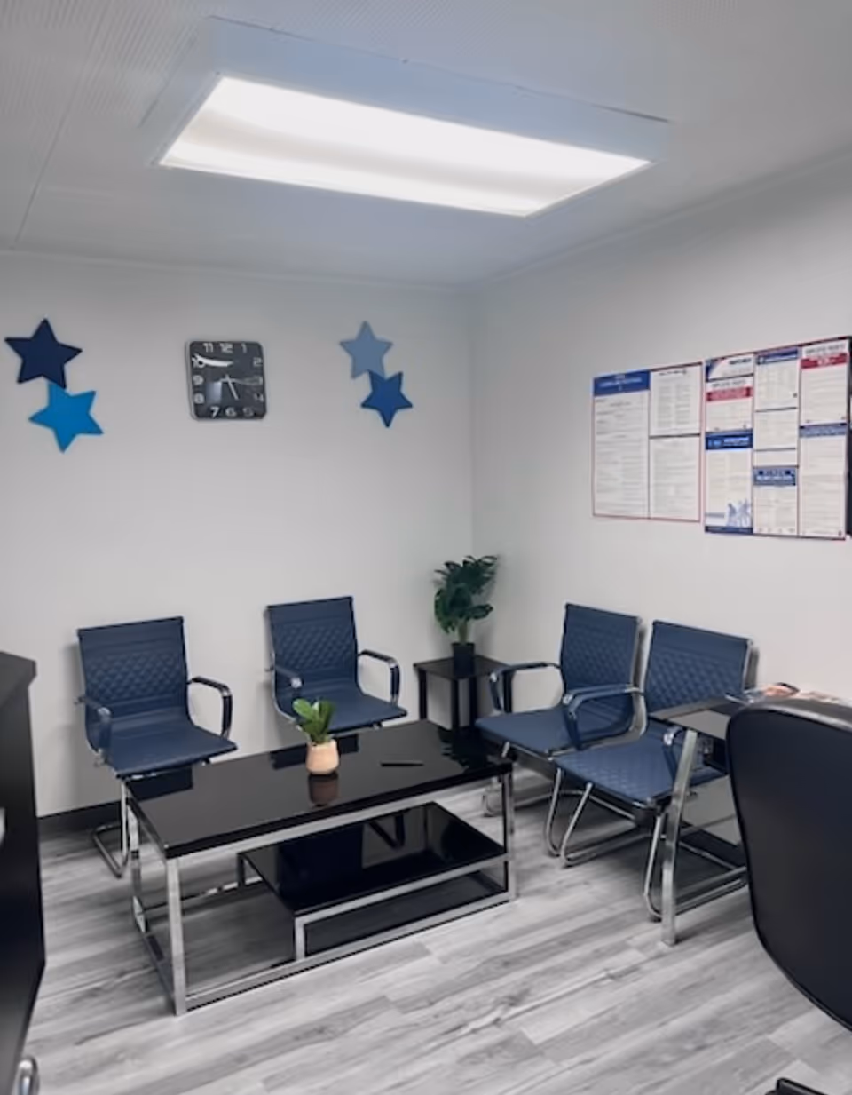Modern waiting area with blue chairs, glass coffee table, clock, plants, and bulletin board.