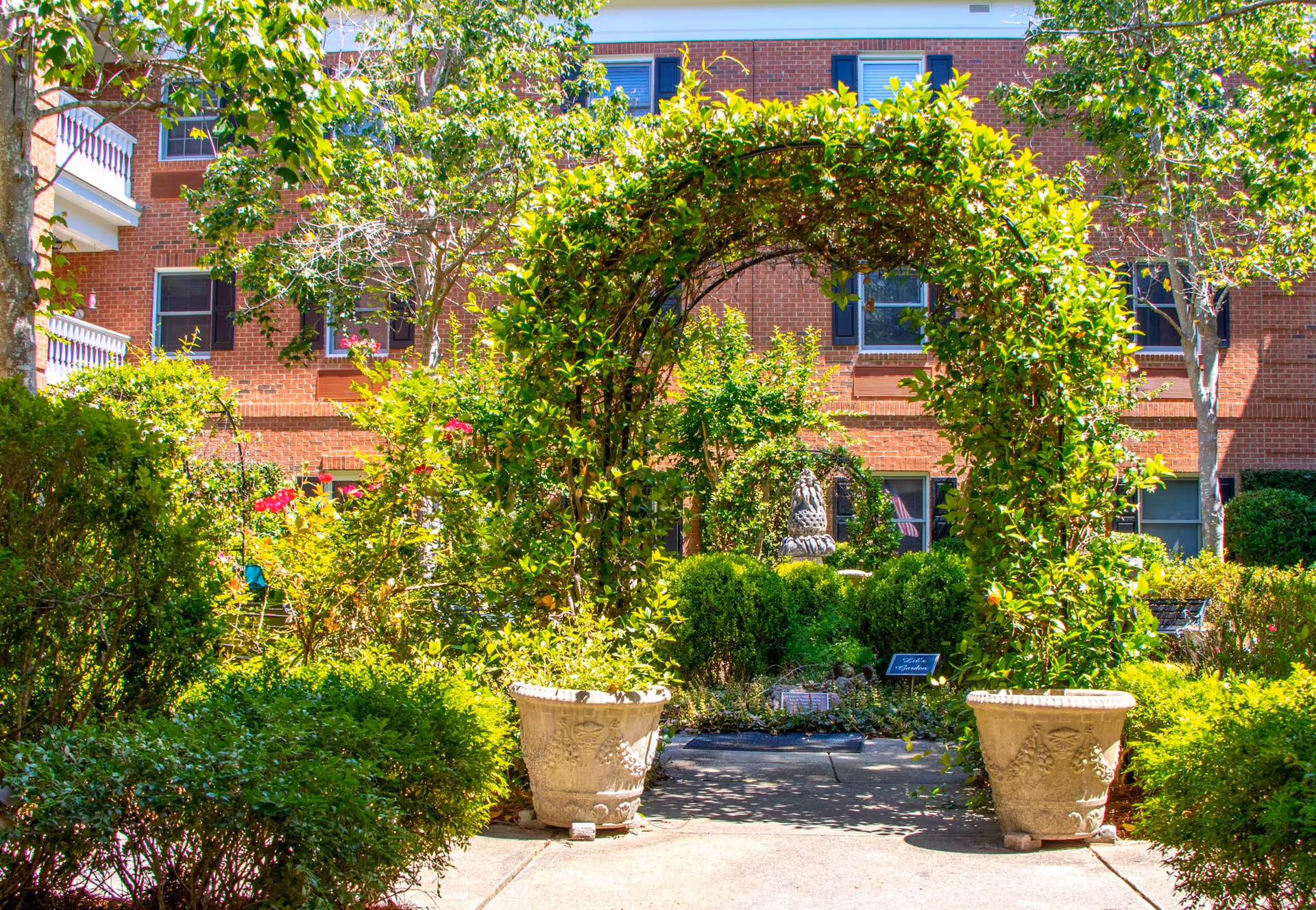 A lush garden area with a green leafy archway supported by two large decorative planters. The garden is filled with various shrubs, bushes, and flowering plants. In the background, there is a red brick building with several windows and balconies.
