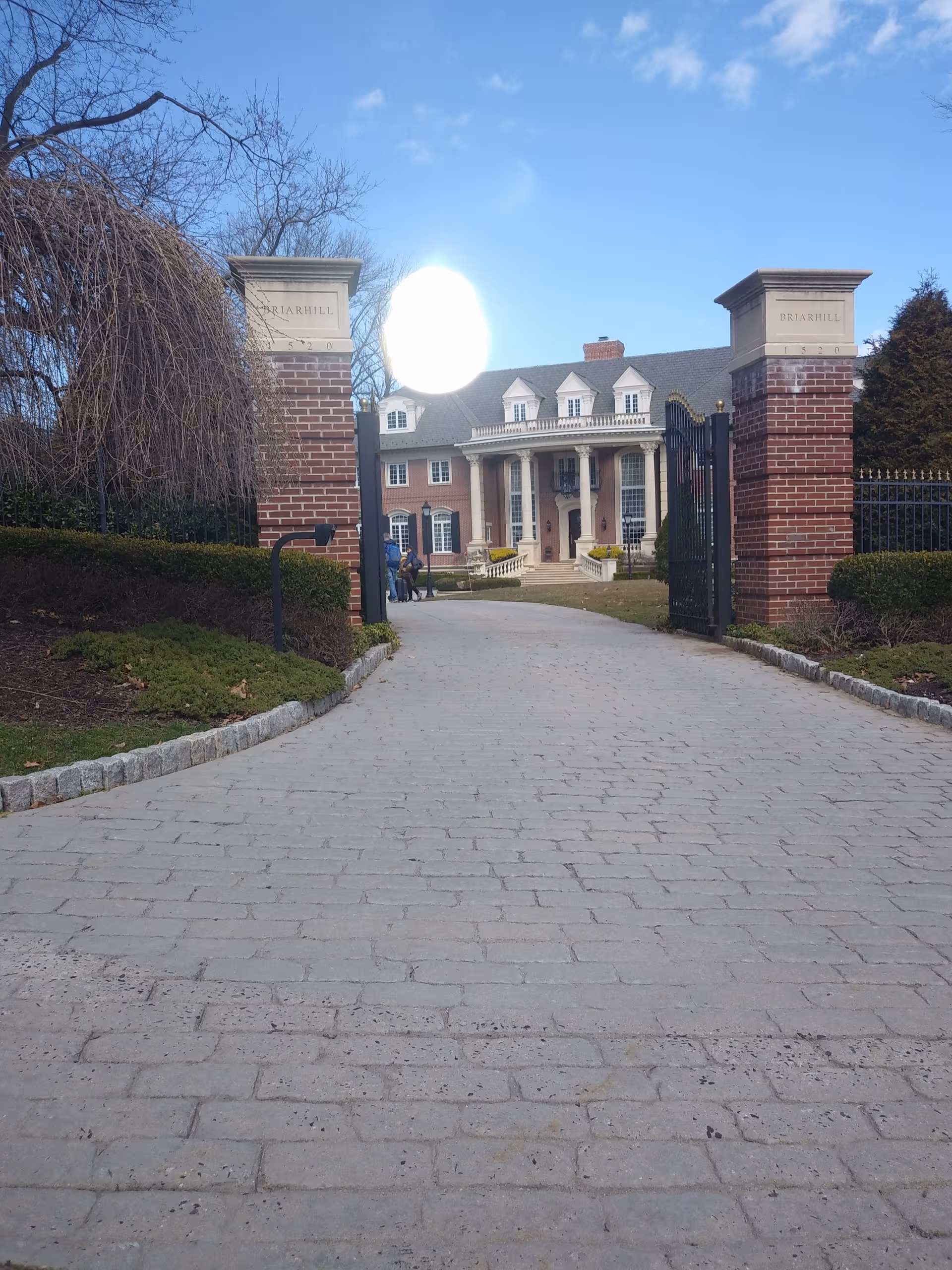 Gated brick entrance leads to a stately building with columns and a cobbled driveway.