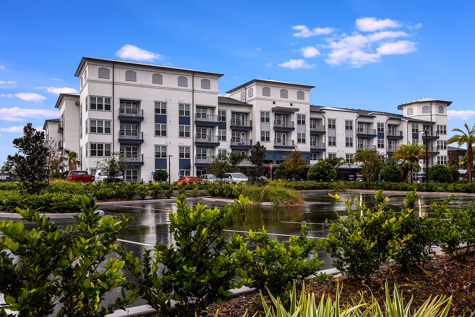 Four-story white apartment-style building with balconies viewed across landscaped grounds and a reflecting pond under a blue sky.