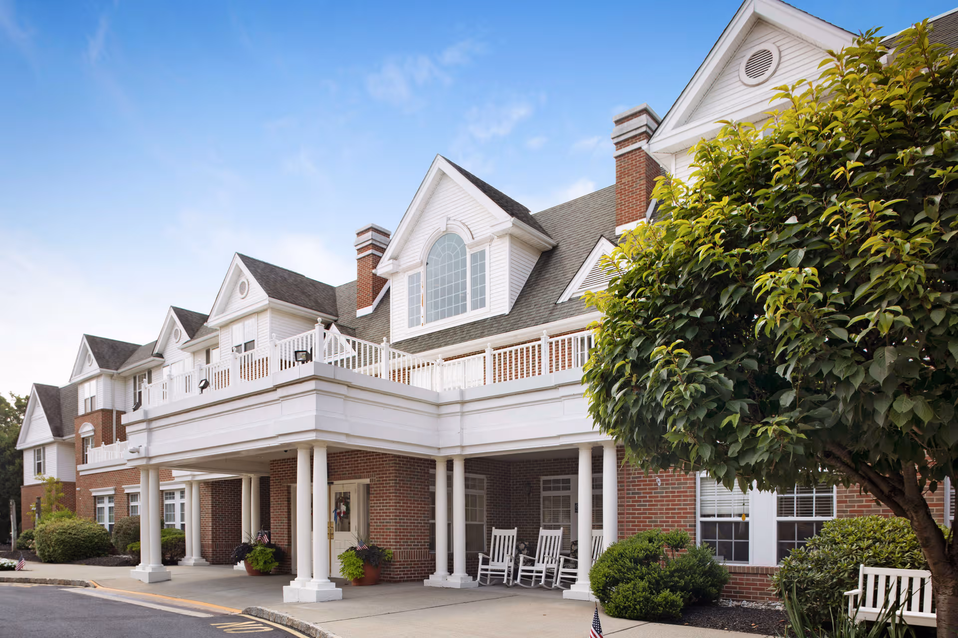 Exterior view of a brick senior living building with a covered entrance and chairs.