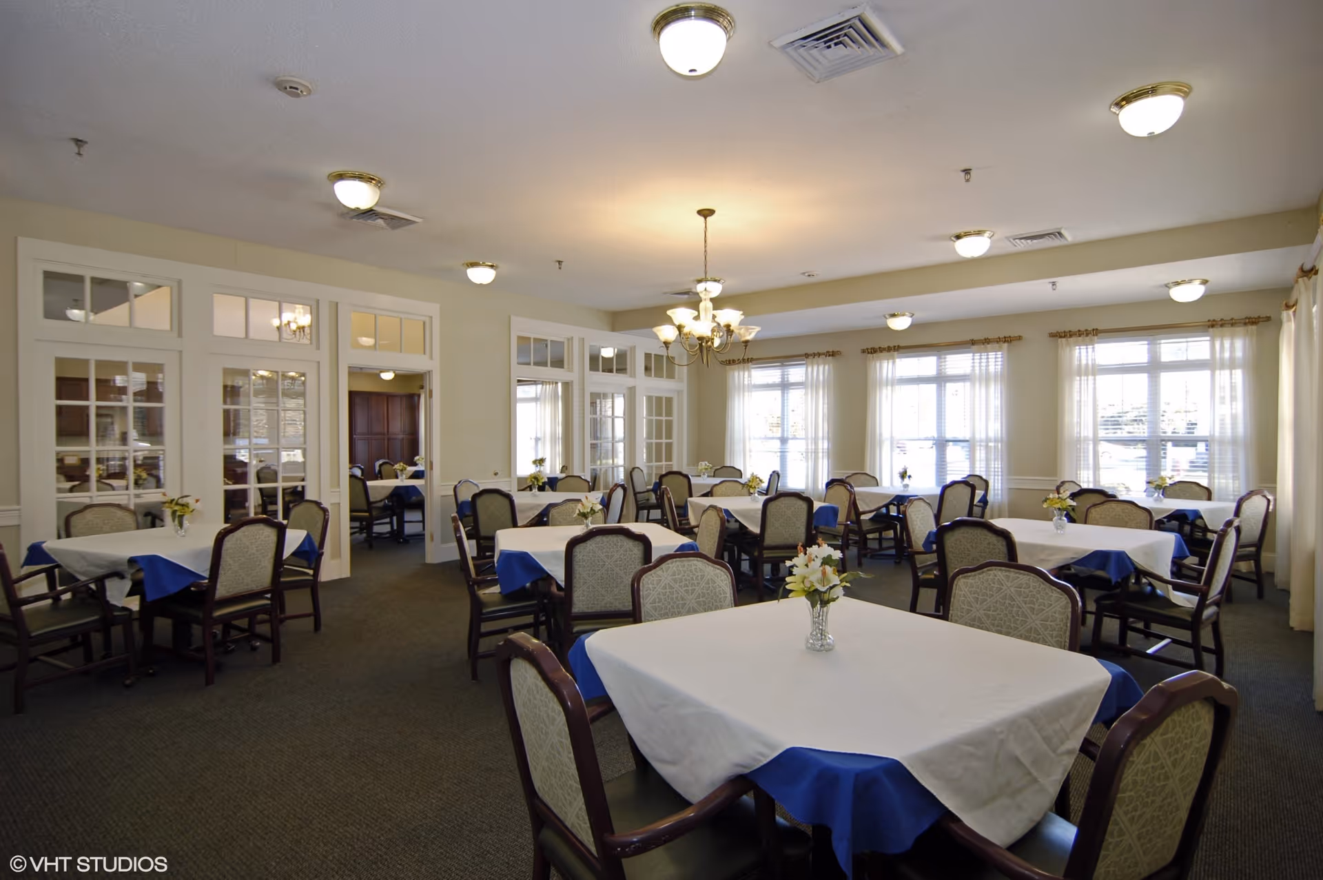 A spacious dining room in a senior living facility with multiple tables covered with white and blue tablecloths, each adorned with a small flower arrangement. The room has large windows with sheer curtains allowing natural light to fill the space. There are upholstered chairs around each table, and the ceiling features several light fixtures including a chandelier.