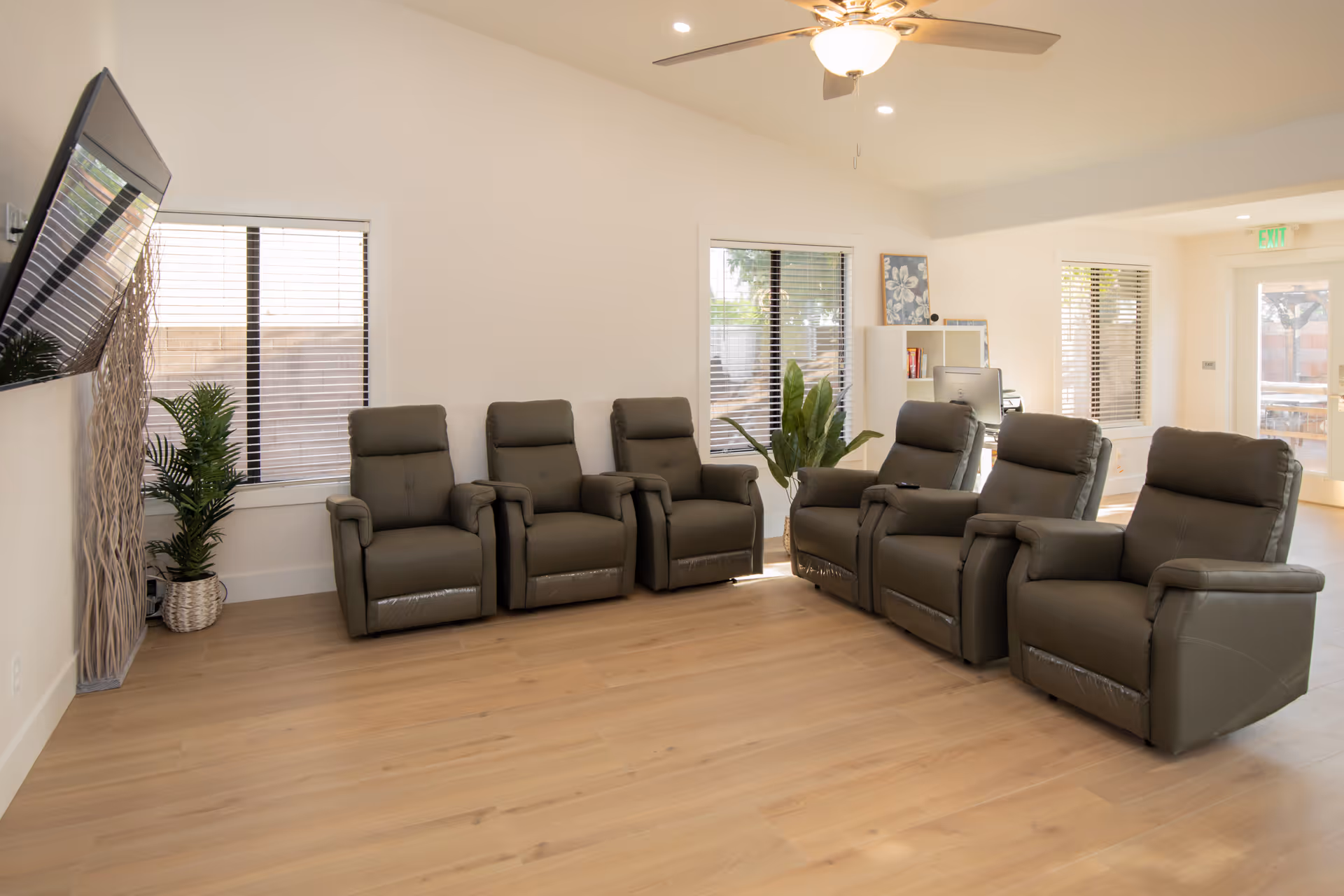 A bright and spacious living room with six gray recliner chairs arranged in two rows facing a wall-mounted flat-screen TV. The room has light wood flooring, white walls, two windows with blinds, a ceiling fan with a light, and some green plants near the windows. There is also a small desk with a computer and bookshelf in the background.