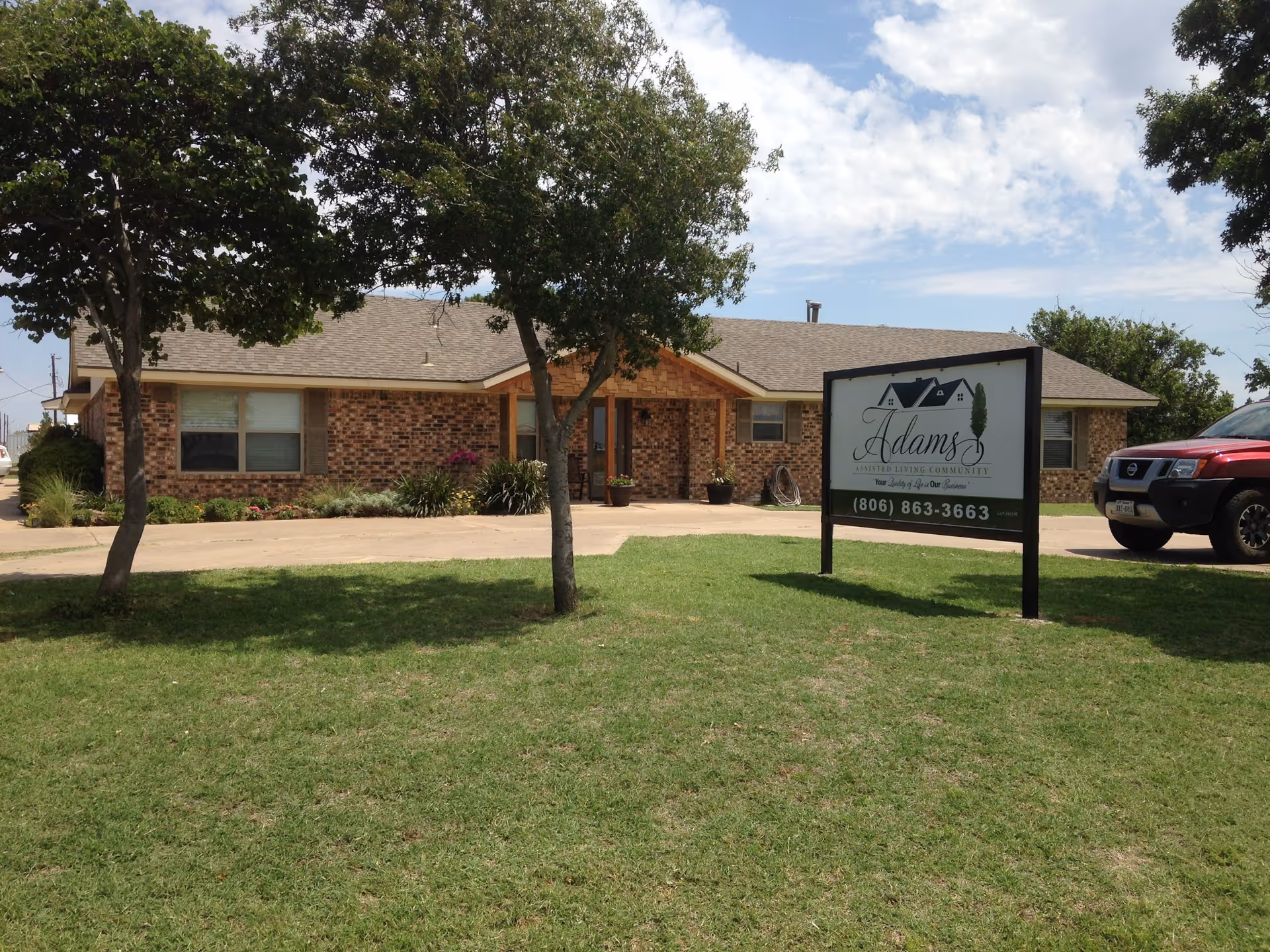 Front view of a single-story brick assisted living building with trees, a lawn, a parked car, and a sign reading 'Adams Assisted Living Community.'