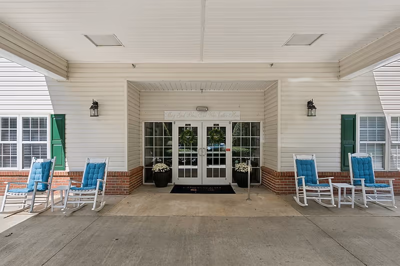 Entrance of Cameron Hall of Canton featuring double glass doors with wreaths, flanked by two large potted plants with white flowers. The building exterior is white with brick accents and green shutters on windows. Four white rocking chairs with blue cushions and two small white tables are arranged on either side of the entrance under a covered porch.
