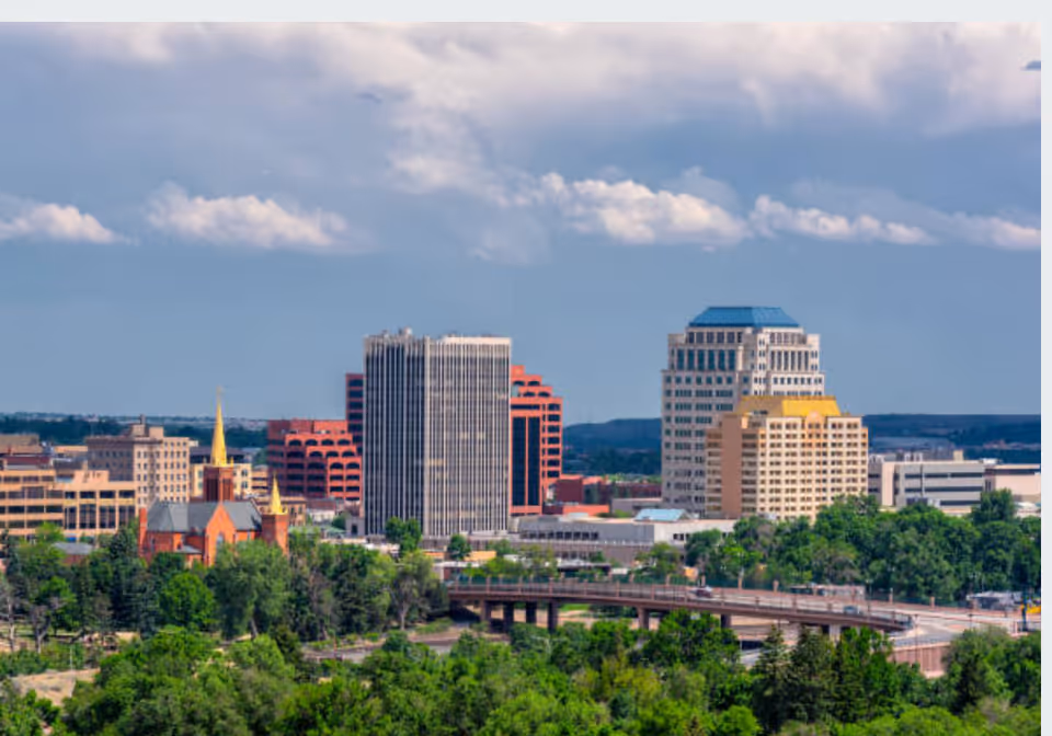 A city skyline with tall buildings, green trees, a highway, and a cloudy sky.