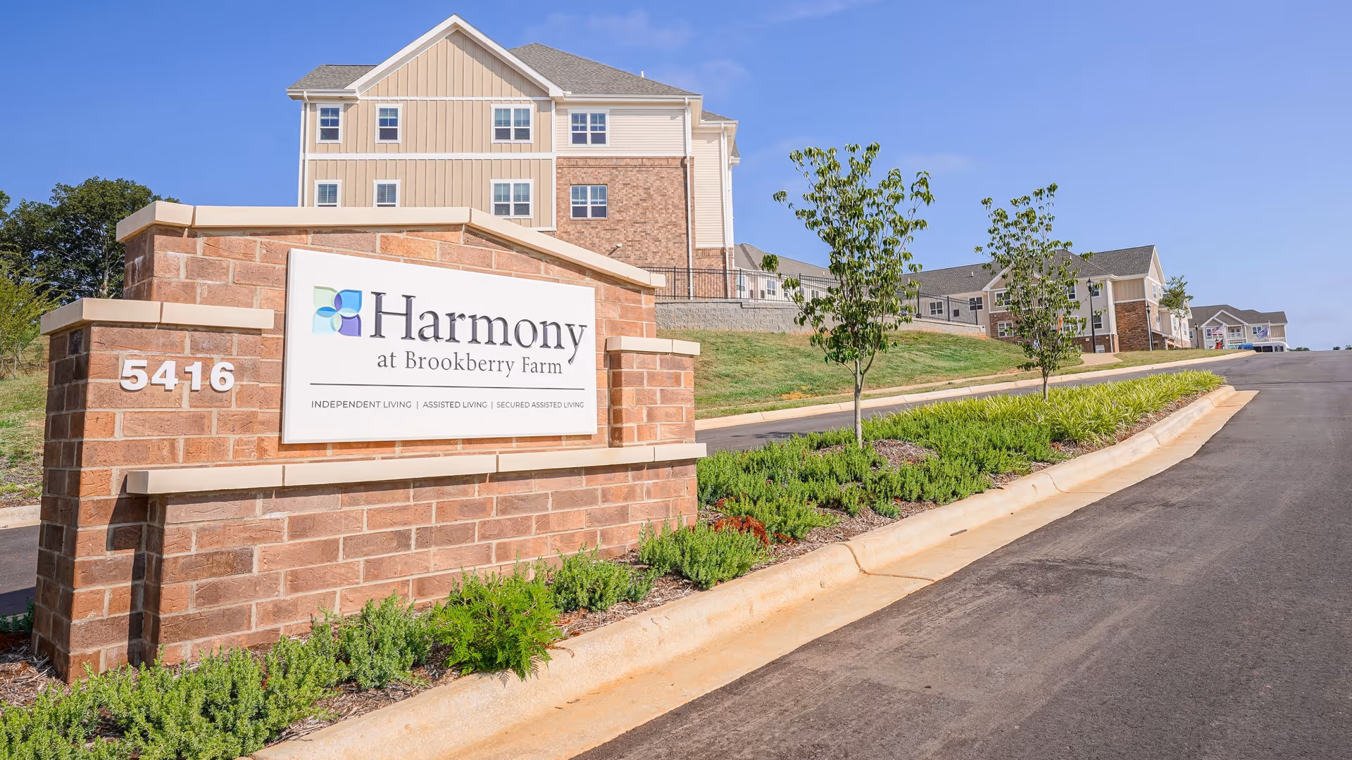 Entrance sign for Harmony at Brookberry Farm senior living facility with a brick base and white sign displaying the facility name and services. Behind the sign are multi-story residential buildings, a paved road, green landscaping, and a clear blue sky.