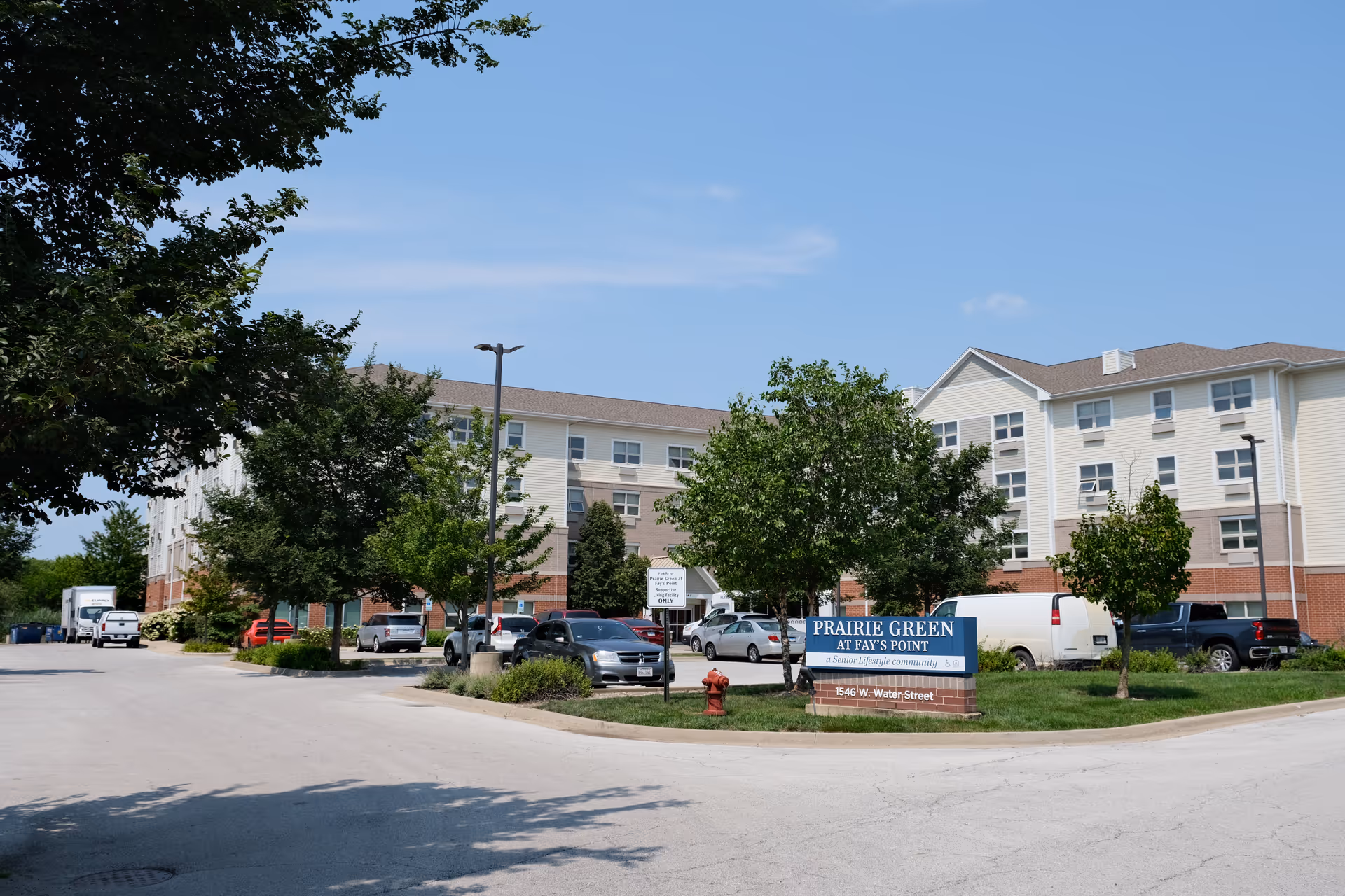 Front exterior of the Prairie Green at Fay's Point senior living building with parking lot, trees, and a facility sign under a blue sky.
