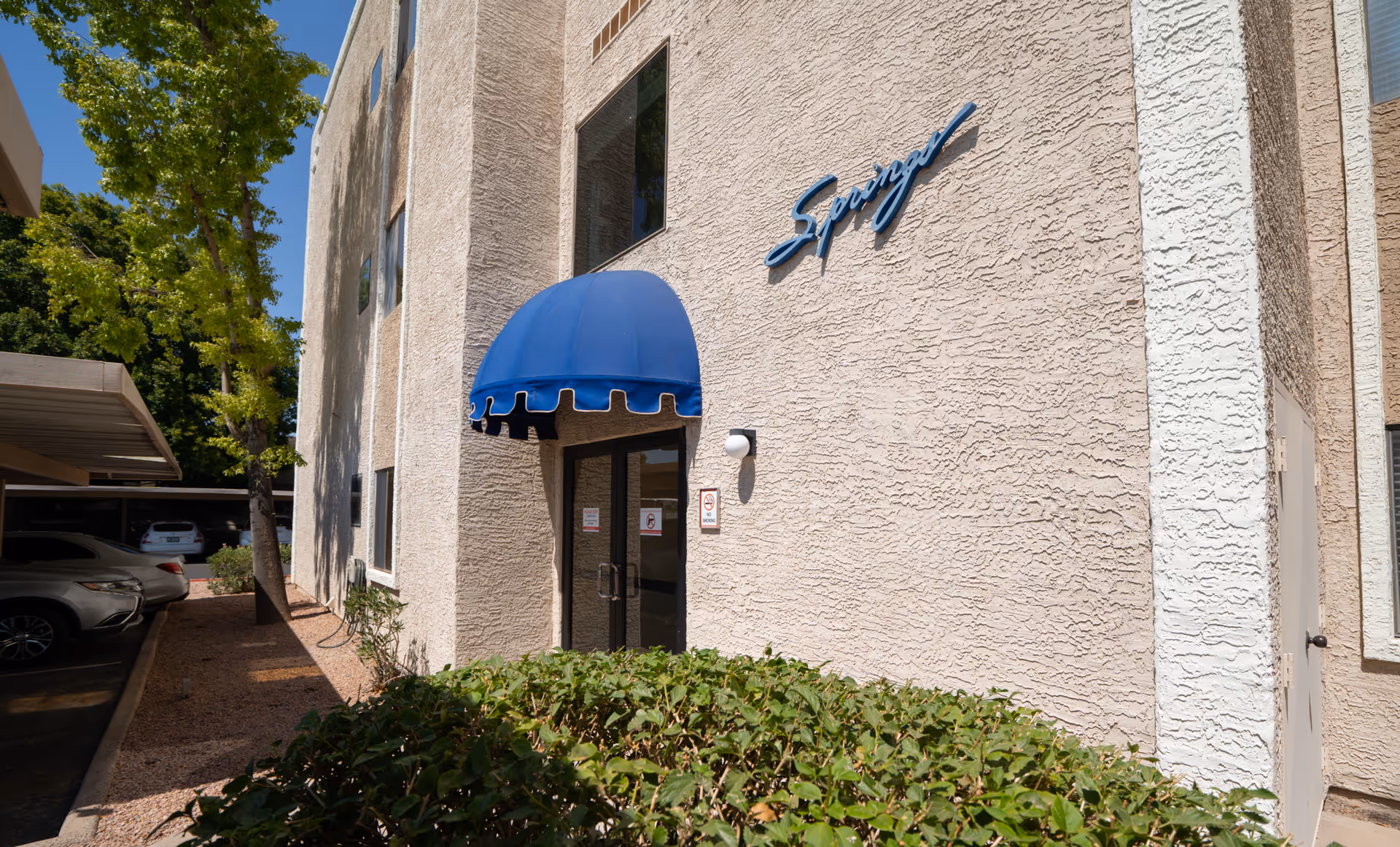 Exterior view of The Springs of Scottsdale entrance with a blue awning and beige textured walls.