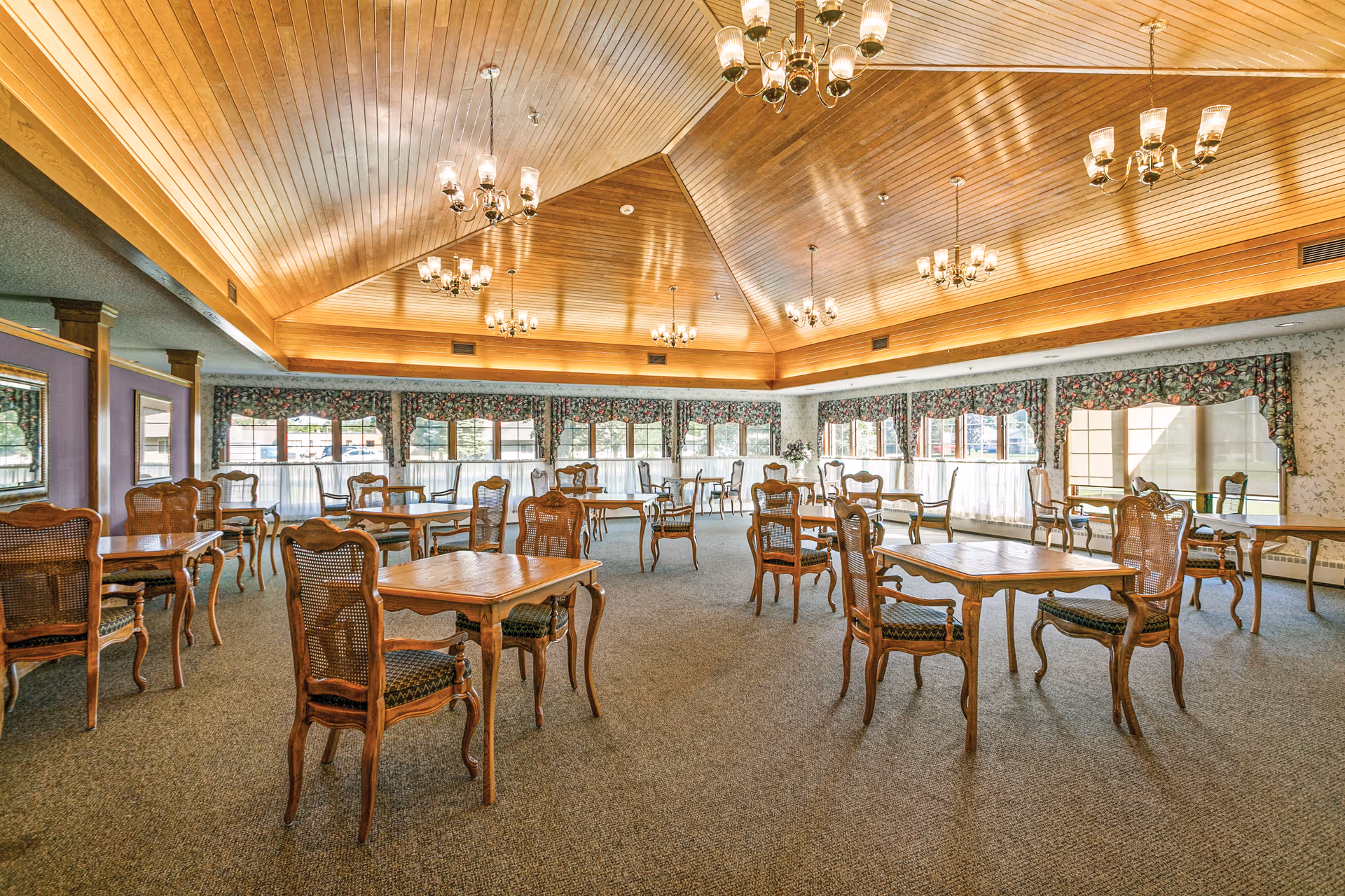 A spacious dining room with multiple wooden tables and chairs arranged neatly on a carpeted floor. The room features a high, wooden vaulted ceiling with several elegant chandeliers providing warm lighting. Large windows with floral curtains allow natural light to fill the space.