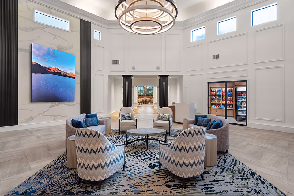 A spacious and modern common area with high ceilings and large windows near the ceiling. The room features a circular seating arrangement with patterned armchairs and sofas around a round coffee table on a decorative rug. A large digital screen displaying a scenic lake and mountain view is mounted on the wall. In the background, there is a small convenience area with shelves stocked with snacks and beverages behind glass doors.