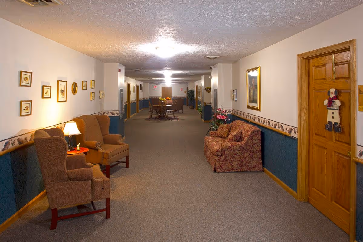 A long, carpeted hallway in a senior living facility with beige walls and blue wainscoting. The hallway is furnished with two brown armchairs and a small table with a lamp on the left side, and a patterned sofa with a floral arrangement on the right side. Several framed pictures and a clock hang on the walls. At the far end of the hallway, there is a table with chairs and some plants. Wooden doors line the hallway, one decorated with a snowman ornament.