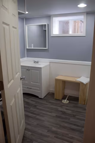 Small bathroom with a vanity, mirror, window, and wooden bench beside a floor drain.