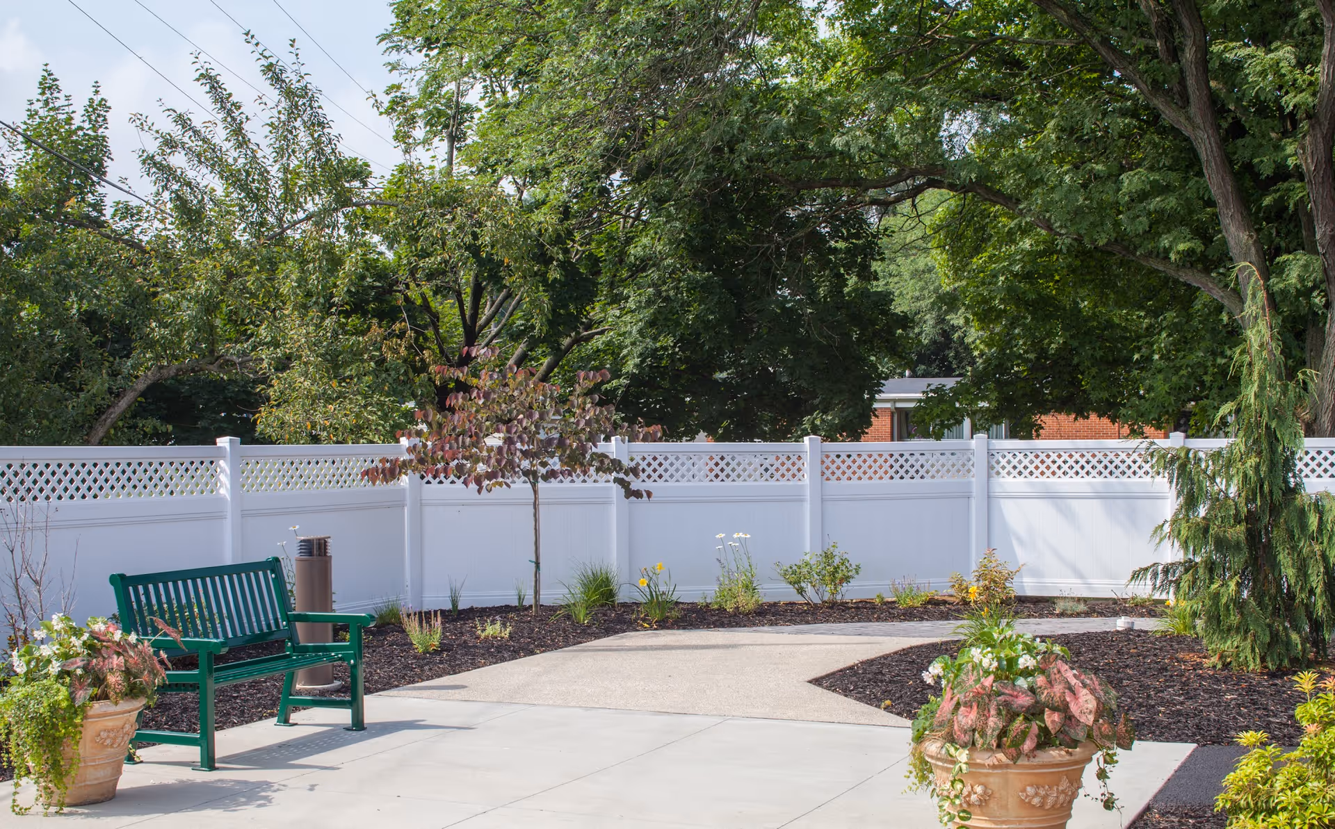 Outdoor garden area with a green bench, potted plants, a white privacy fence, and various trees and shrubs under a partly cloudy sky.