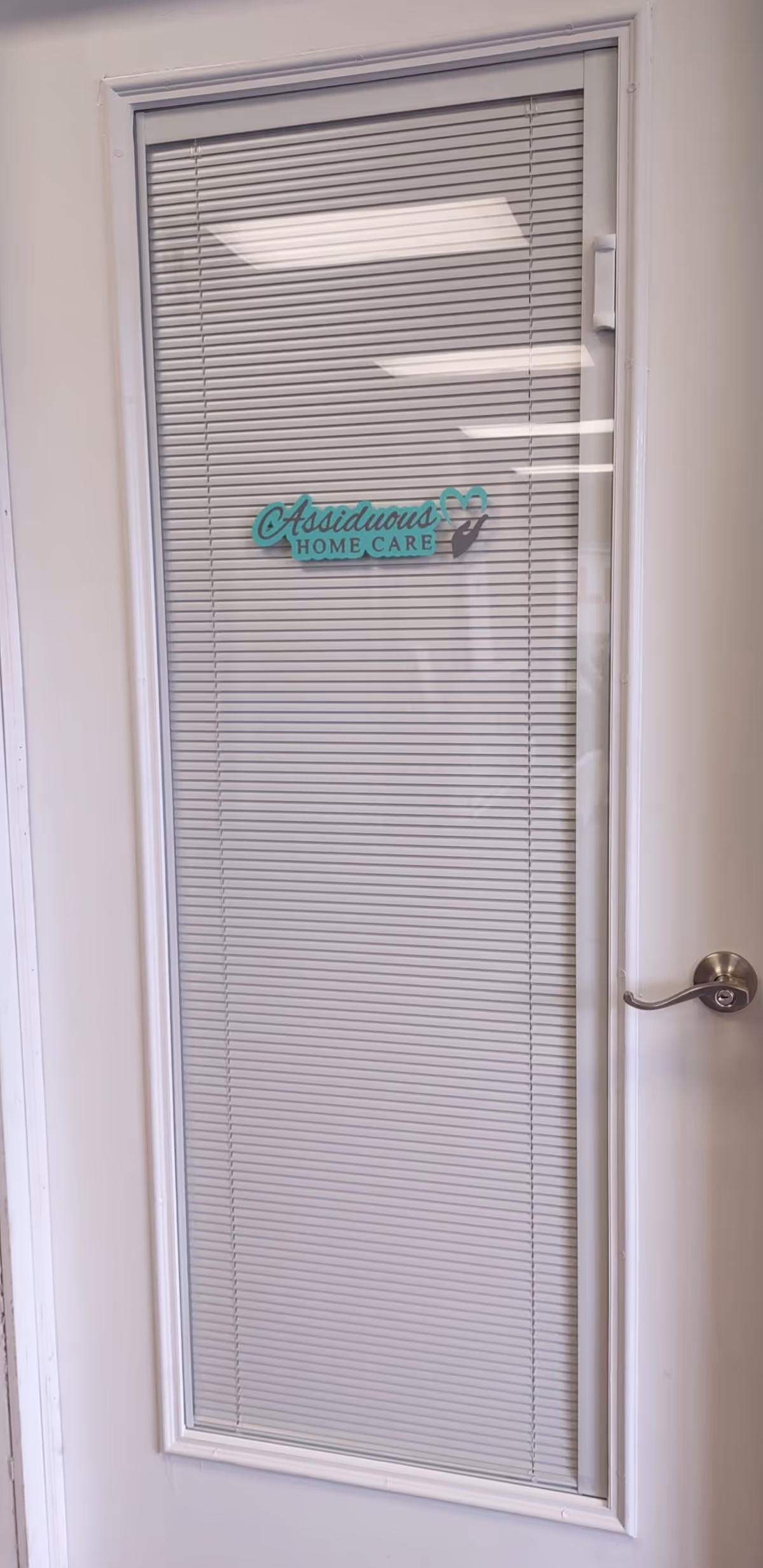 A glass door with blinds displays the Assiduous Home Care logo.