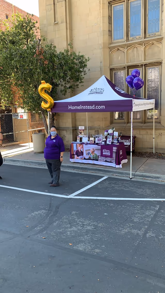 Purple Home Instead tent outside a building with brochures and balloons, staffed by a caregiver.
