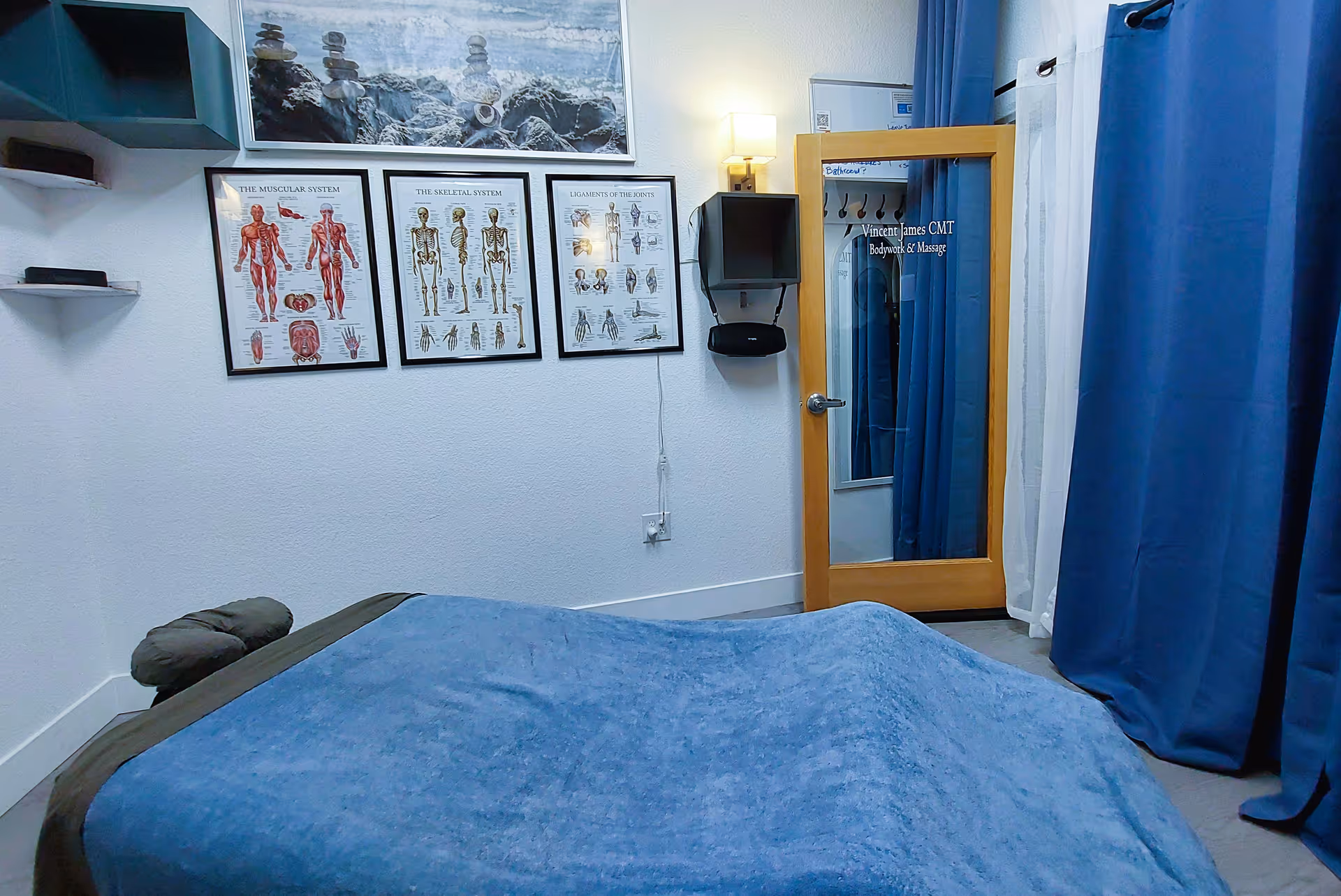 Massage room with a blue-covered table, medical posters, and a doorway labeled Vincent James CMT.
