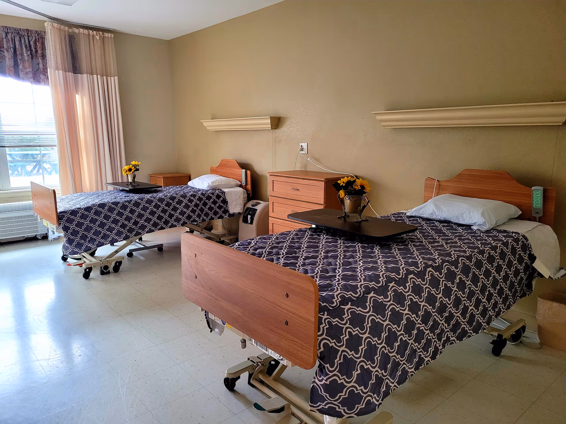 A bright shared patient room with two hospital-style beds, patterned bedspreads, overbed tables, nightstands, and a window with curtains.