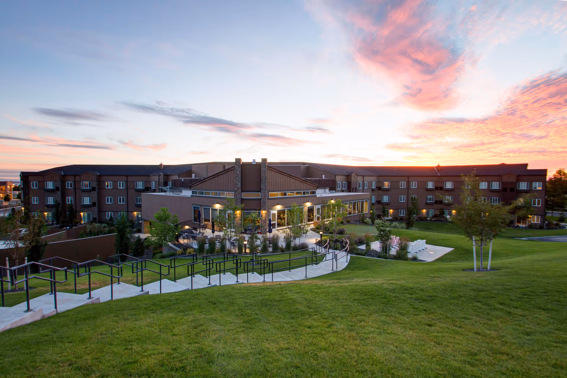 Wide exterior view of a large senior living facility building at sunset with a well-maintained green lawn, trees, and a paved walkway with handrails leading up to the entrance area.