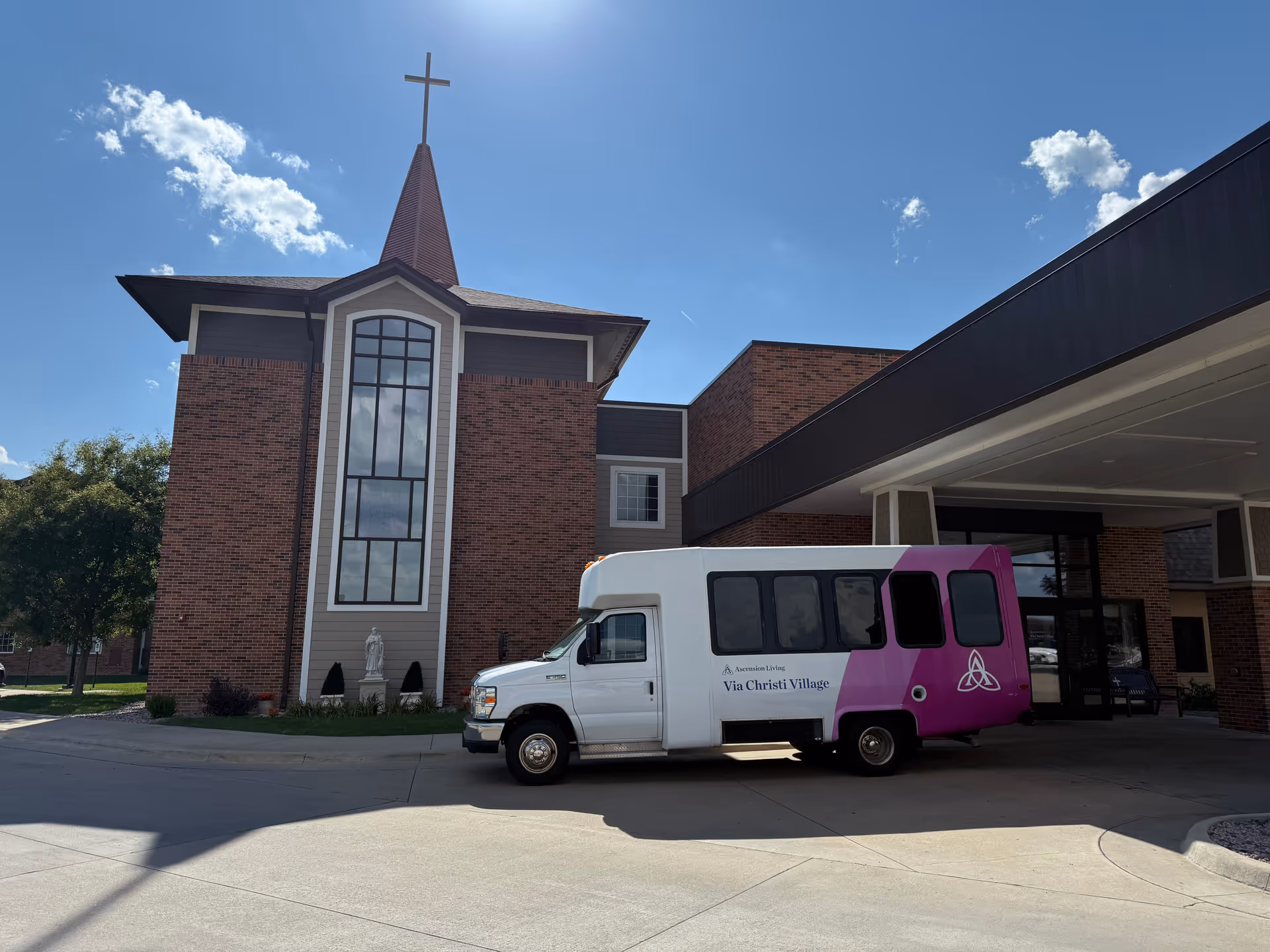 Exterior view of Ascension Living Via Christi Village with a white and pink shuttle bus.