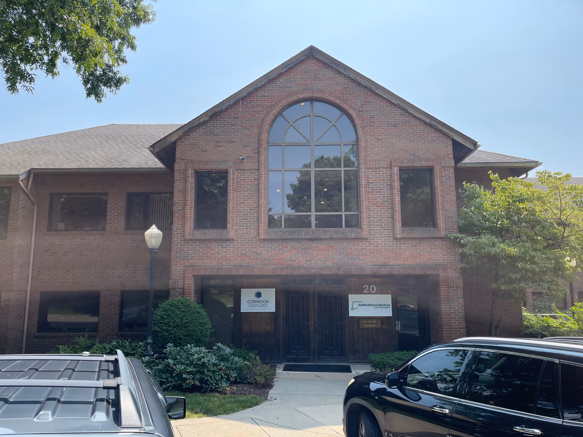 Brick two-story building with a large arched window, entrance doors, and parked cars.