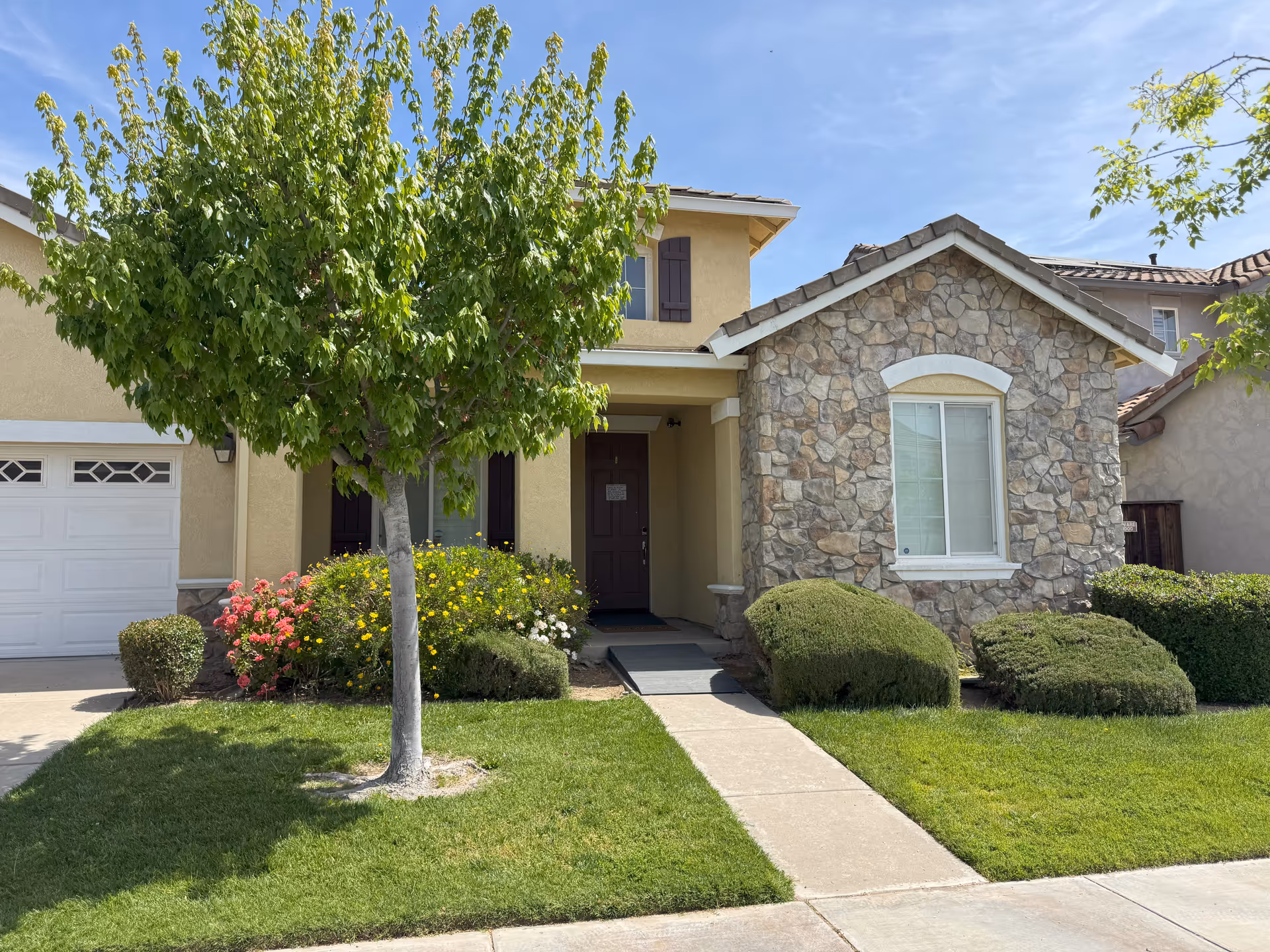 Residential-style assisted living building exterior with front entrance, tree, and manicured shrubs.
