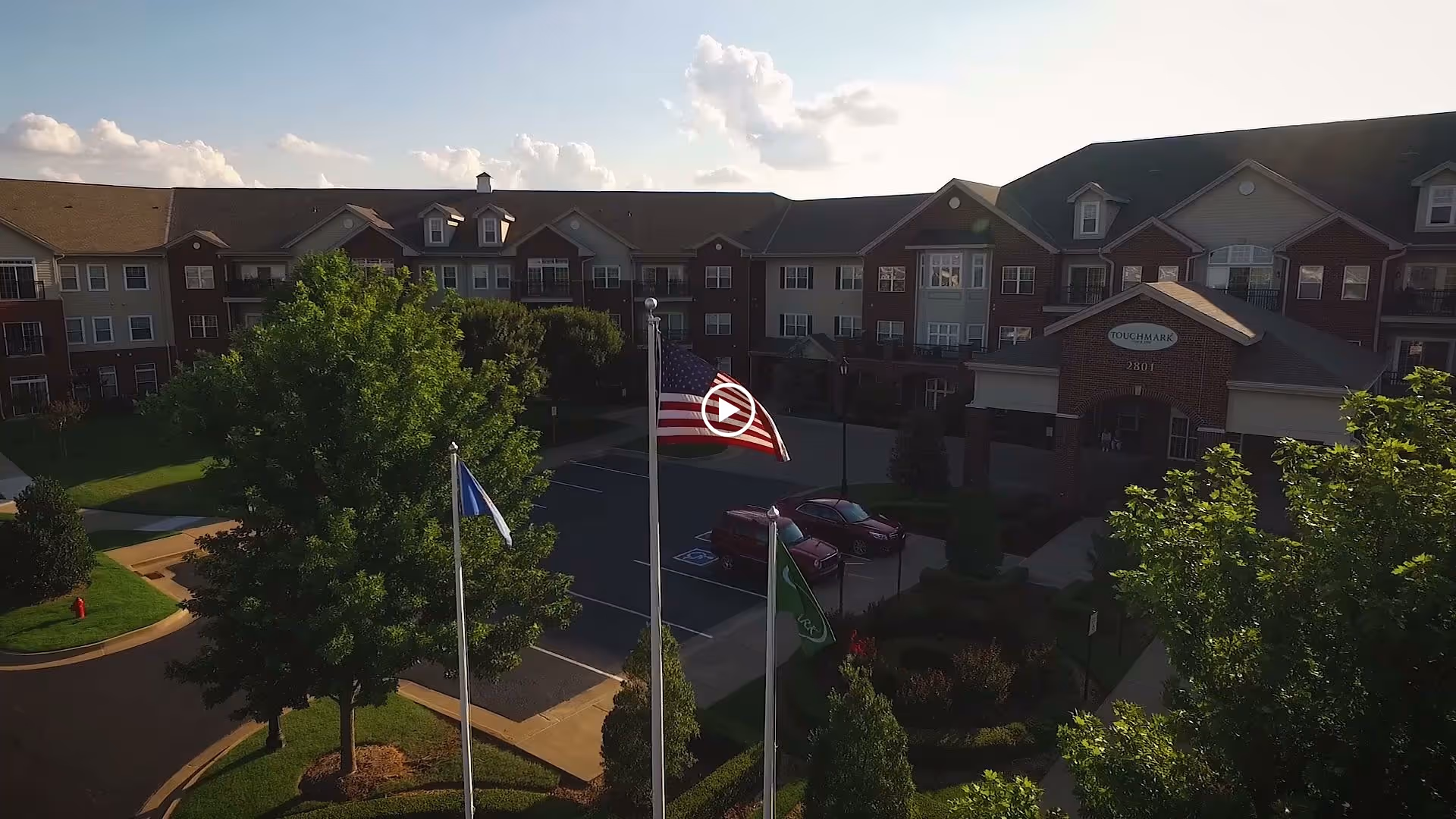 Exterior view of a multi-story senior living facility building with a brick and siding facade, surrounded by trees and a parking lot with several cars. Three flagpoles with flags are visible in the foreground, and the building entrance has a sign that reads 'Touchmark'.