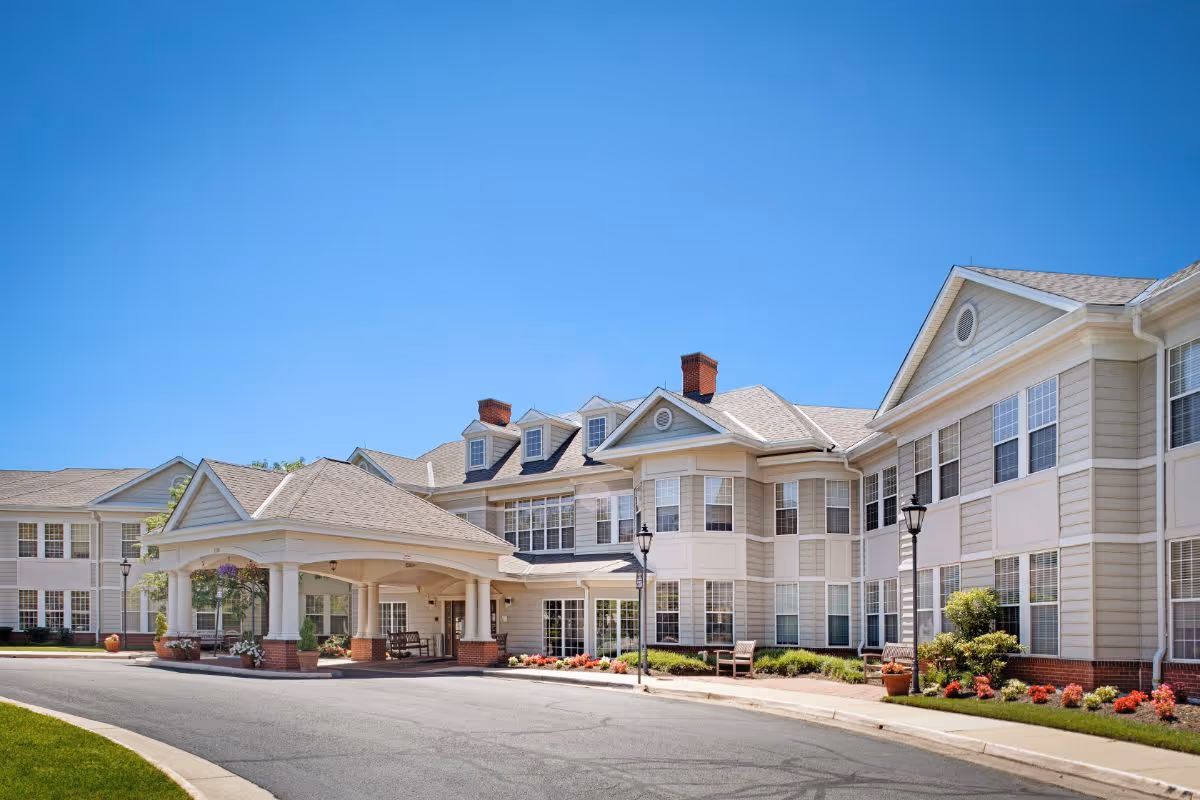 Front exterior of a large multi-story senior living facility with a covered entrance, landscaped beds, and a clear blue sky.