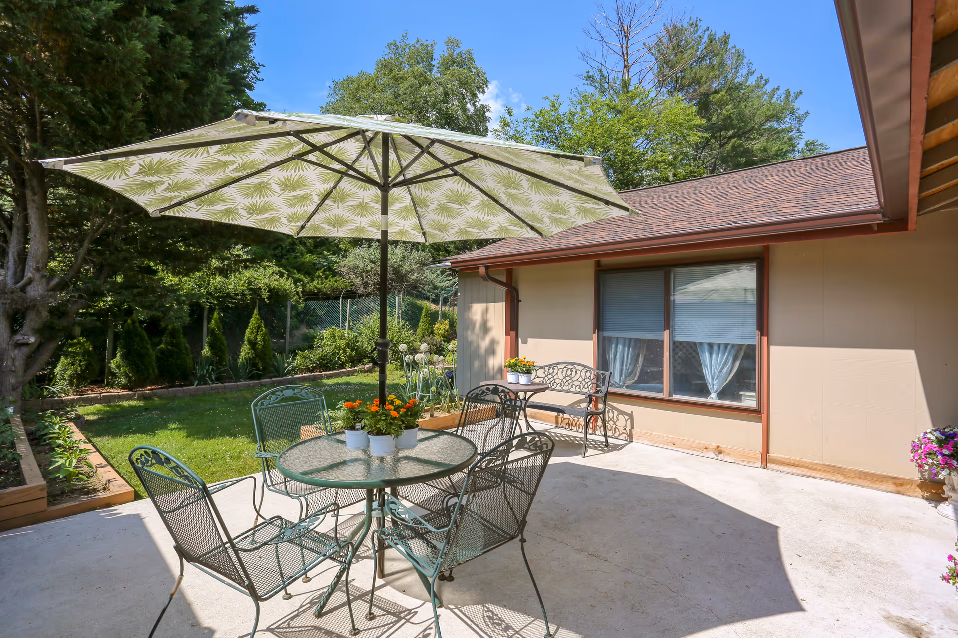 Outdoor patio area with a round glass table, four metal chairs, and a large umbrella with a leaf pattern providing shade. There is a metal bench against the beige exterior wall of the building, with potted flowers on the table and bench. The patio is surrounded by greenery including trees, bushes, and a lawn under a clear blue sky.