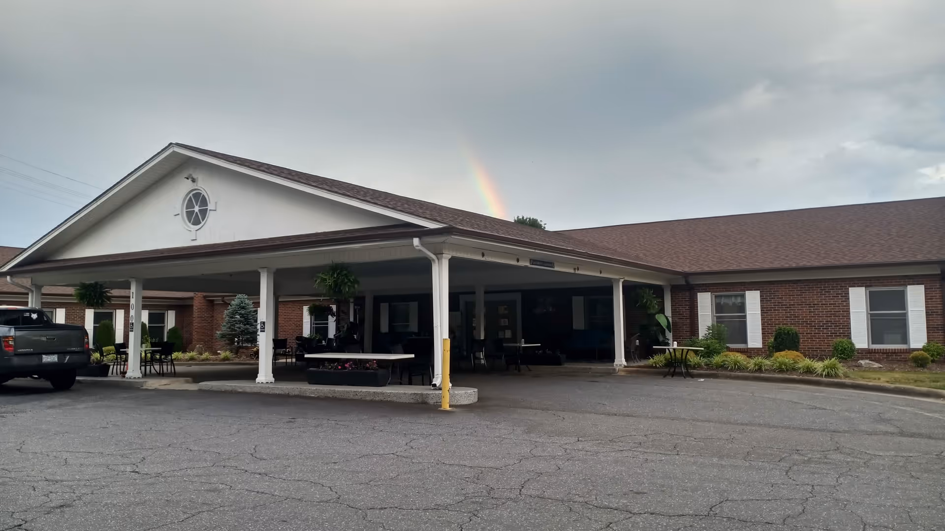 Brick facility with a covered entrance, parking lot, and a rainbow in cloudy sky.