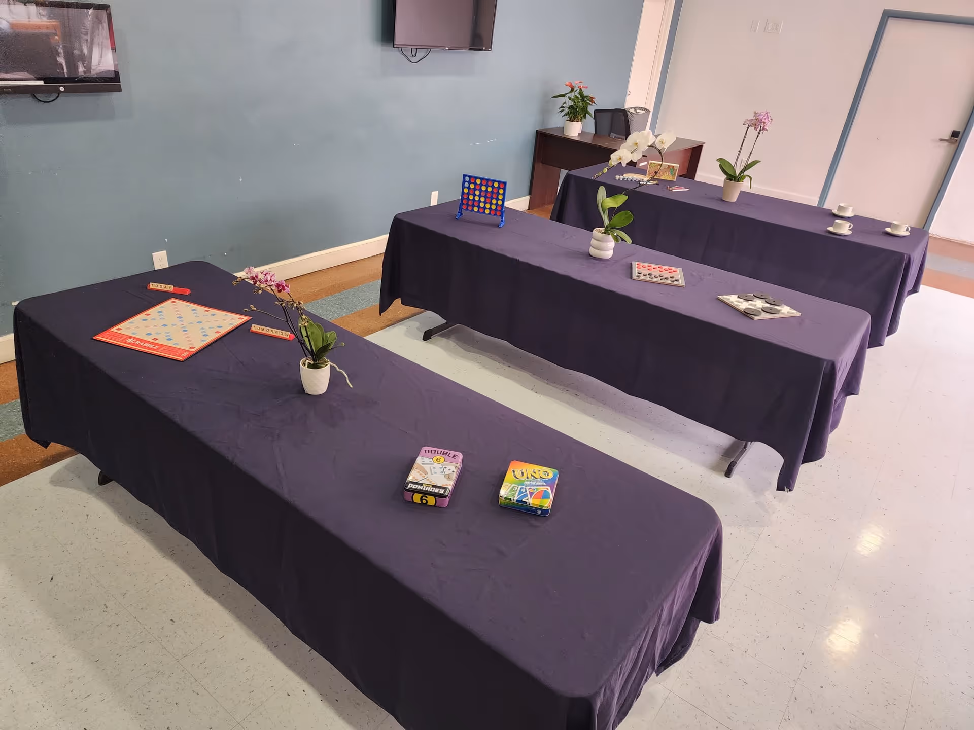 Interior activity room with purple-clothed tables, plants, and board games.