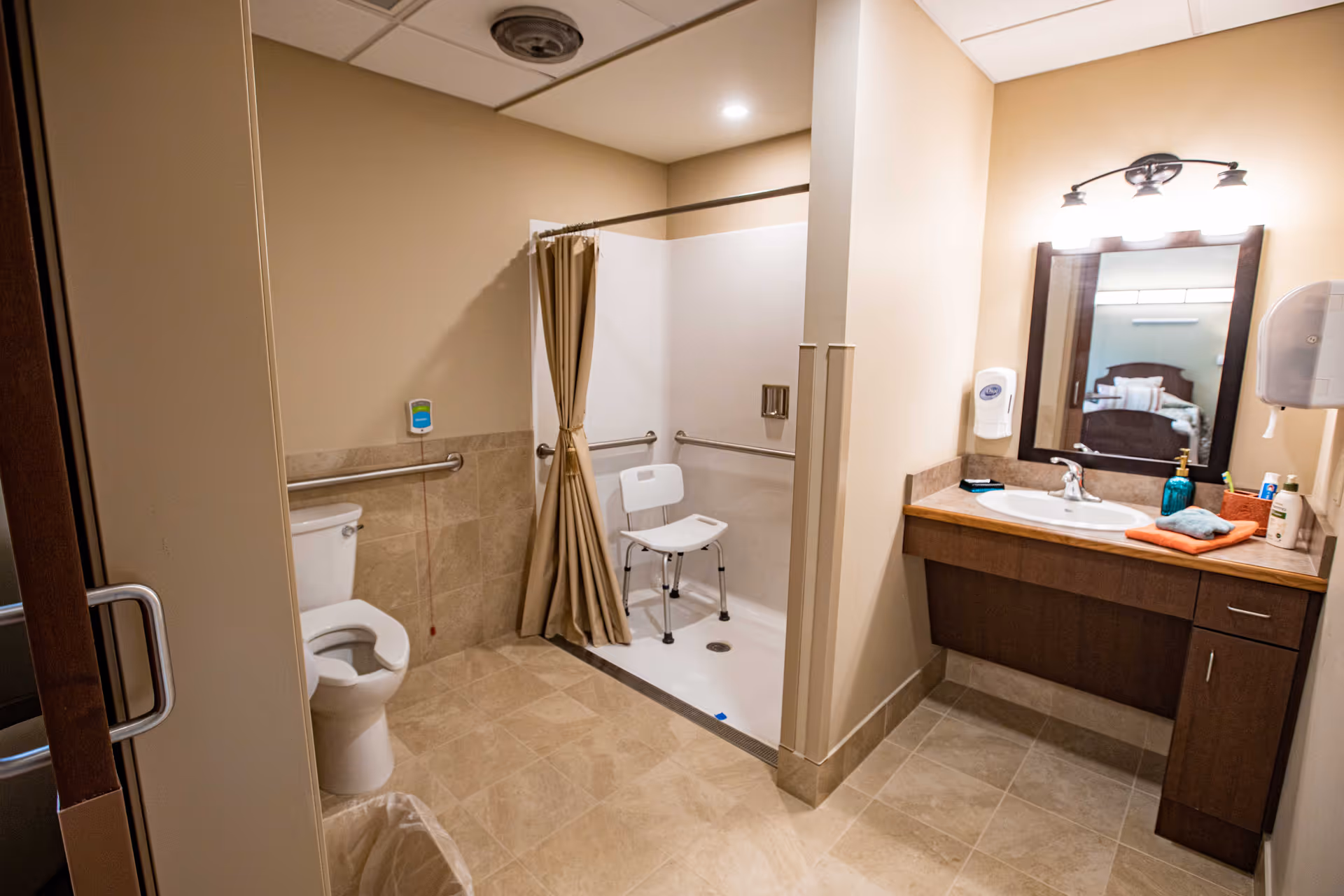 A clean and accessible bathroom with beige tiled floor and walls. The room features a toilet with grab bars, a walk-in shower with a shower chair and curtain, and a wooden vanity with a sink, mirror, and overhead lighting. Various toiletries and a hand soap dispenser are visible on the vanity.