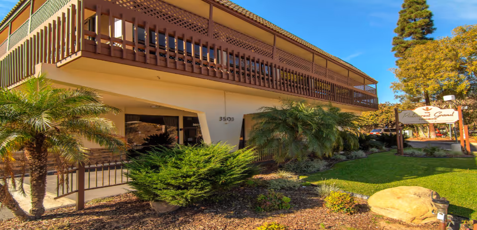 Two-story senior living facility with brown balcony, landscaped yard, and entrance sign.