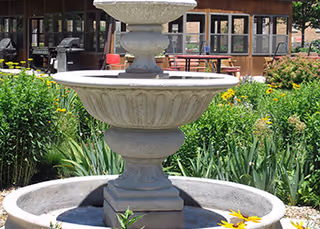 Stone tiered fountain in a garden courtyard with greenery and a building in the background.