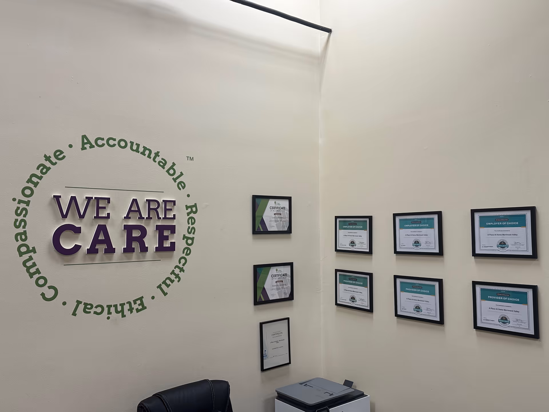 Office corner with wall logo 'We Are CARE' and framed certificates on light walls.