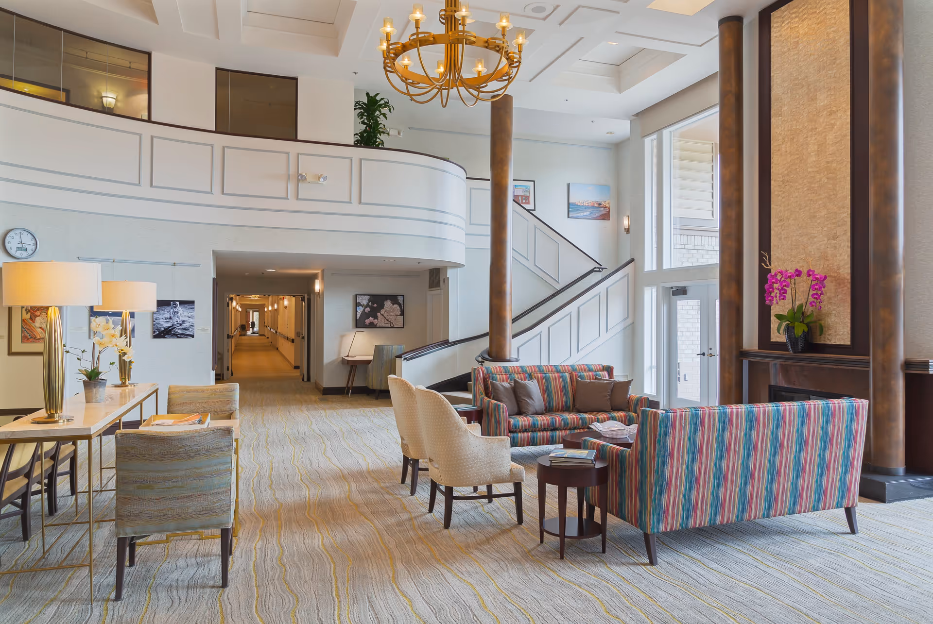 A spacious and well-lit common area in a senior living facility featuring a seating arrangement with two striped sofas and two beige armchairs around a small round table. The room has a high ceiling with a large chandelier, a staircase with a white railing, and large windows letting in natural light. There are decorative plants, framed pictures on the walls, and a long hallway visible in the background.