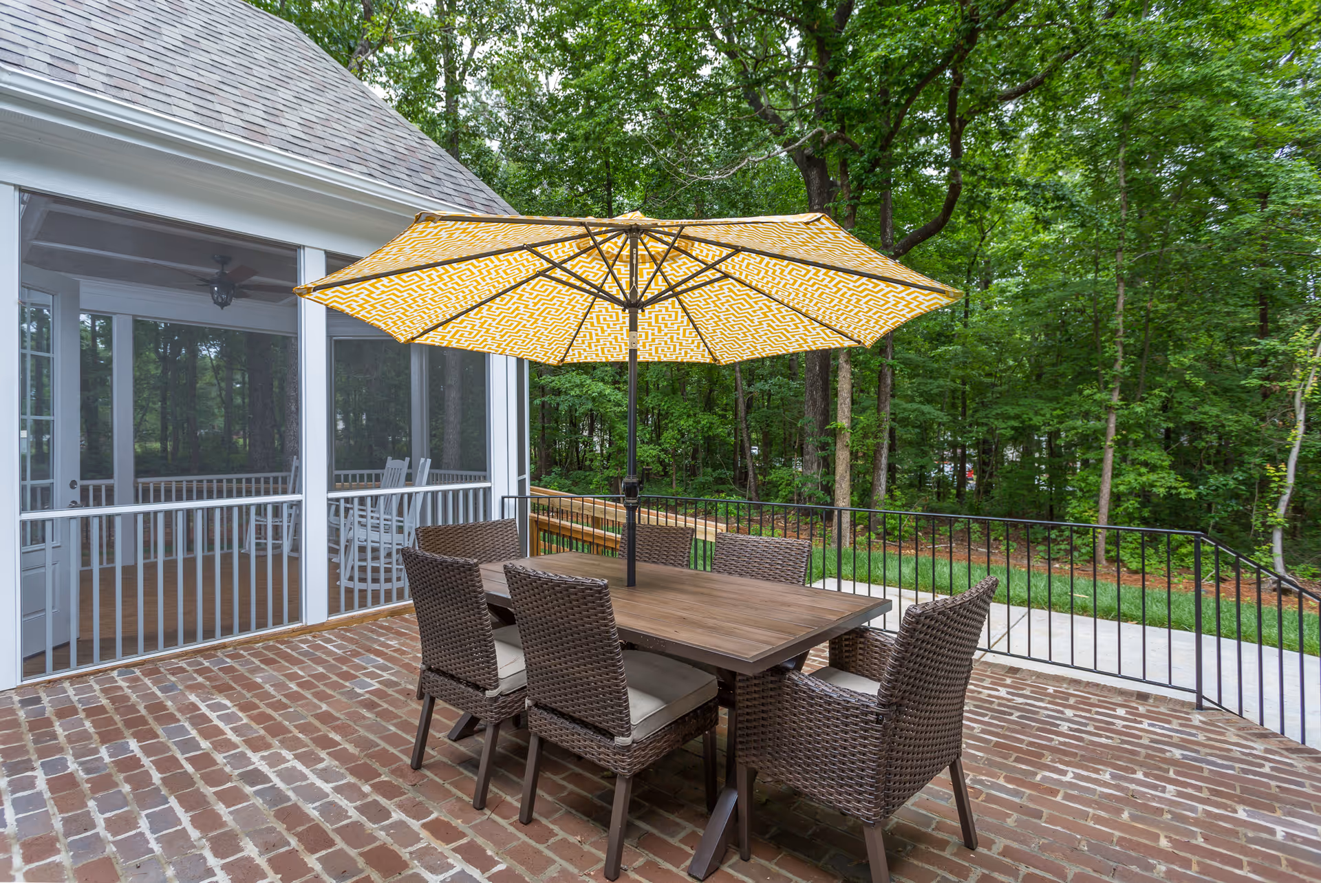 Outdoor patio area with a rectangular wooden table surrounded by six wicker chairs with cushions. A large yellow patterned umbrella is open above the table. The patio has a brick floor and is adjacent to a screened porch with white railings. In the background, there are green trees and a grassy area.